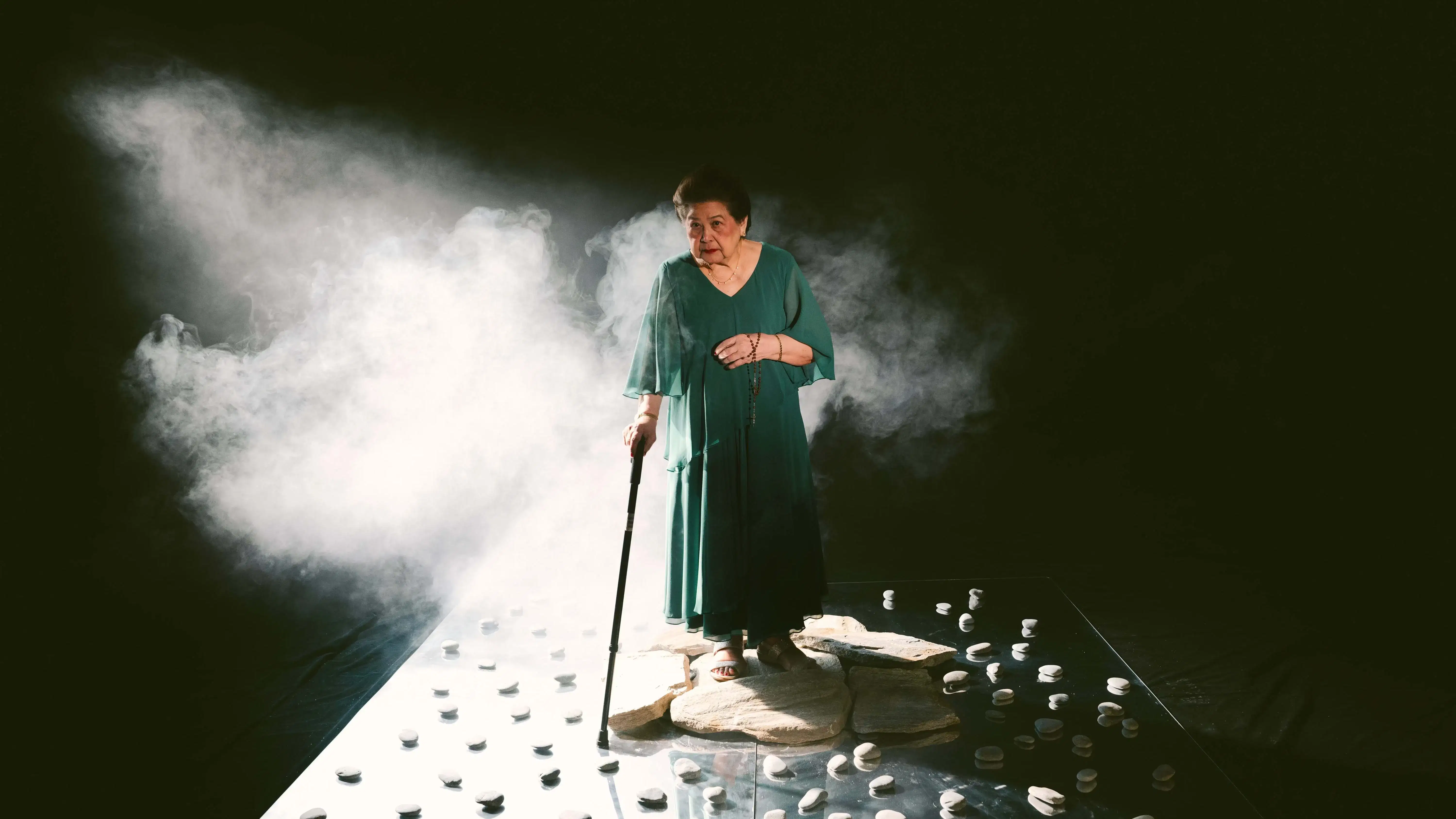 A still from a color video: An elderly woman in a teal dress stands on a cluster of flat stones, holding a cane in her right hand and a rosary in her left. She is surrounded by smoke and placed on a glossy black platform scattered with smooth white stones. The background is dark, with light focused on the figure from the left.