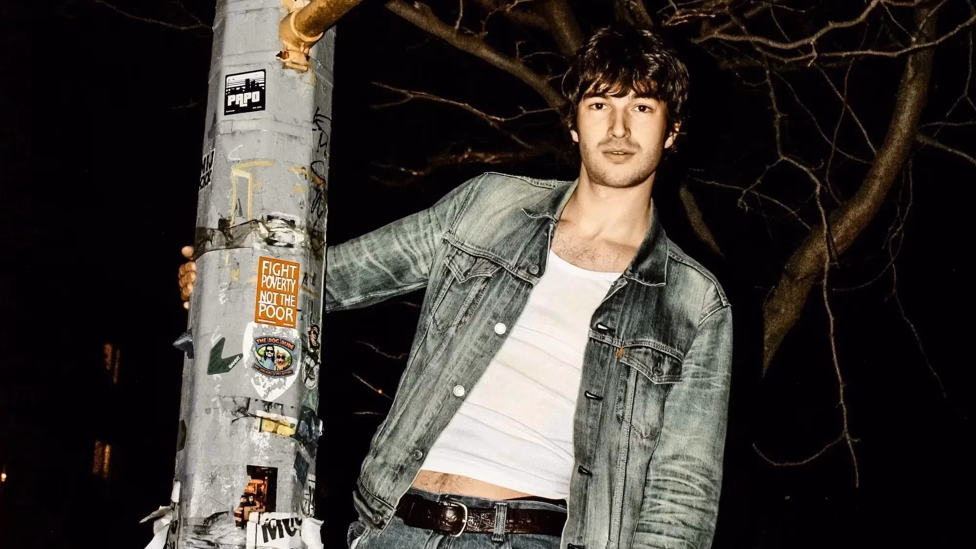 A portrait of man stood on top of, and hanging onto a streetlight at night.