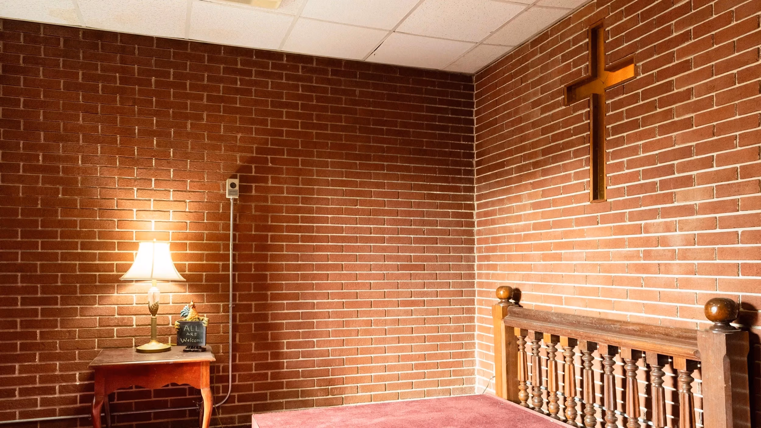 A small prayer room with an altar, side table, and lamp. A cross is embedded in the brick wall.