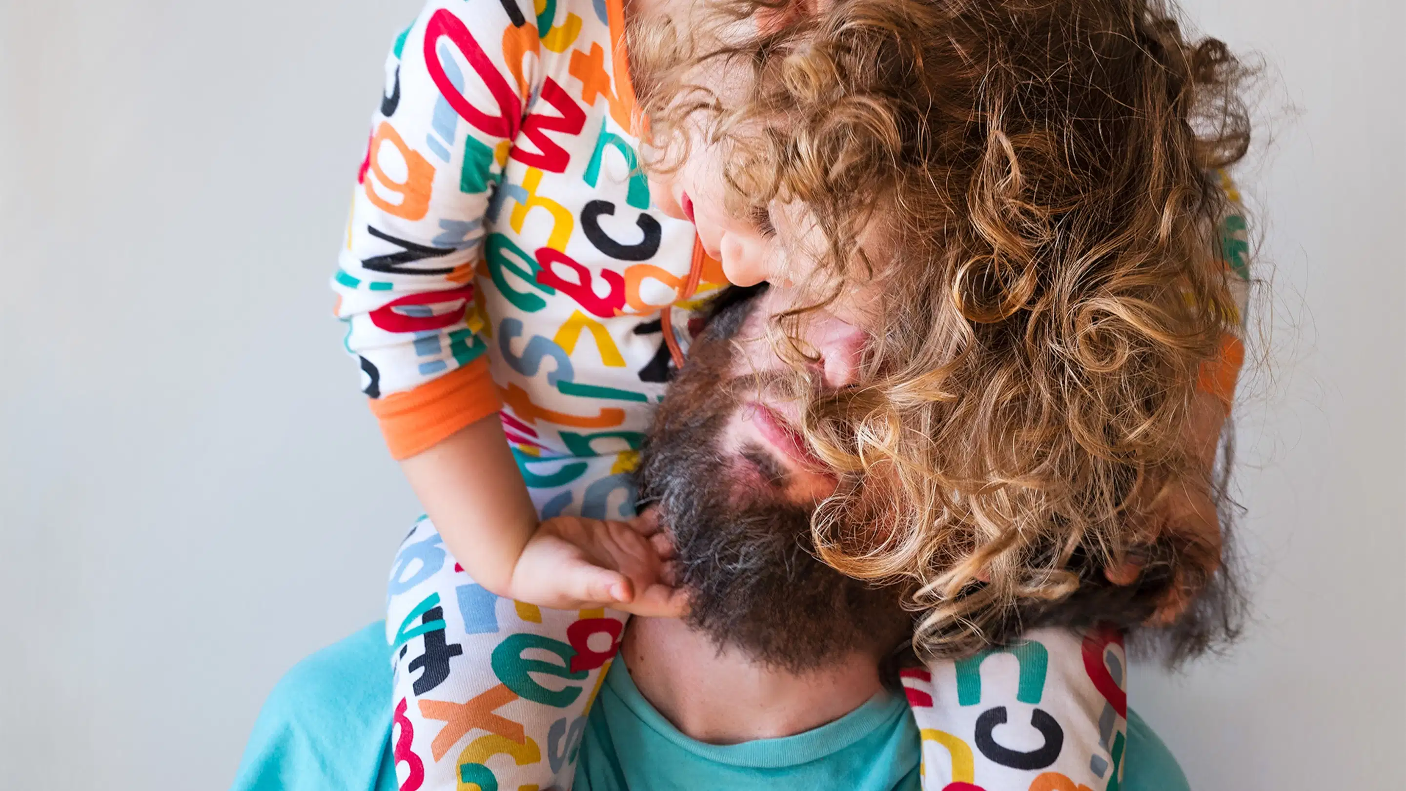 A documentary photograph of a child in a onesie patterned with letters sitting on the shoulders of their father. The child is draped over their head so the father’s eyes are covered. The father is holding their child’s legs and is wearing a blue short sleeve shirt.