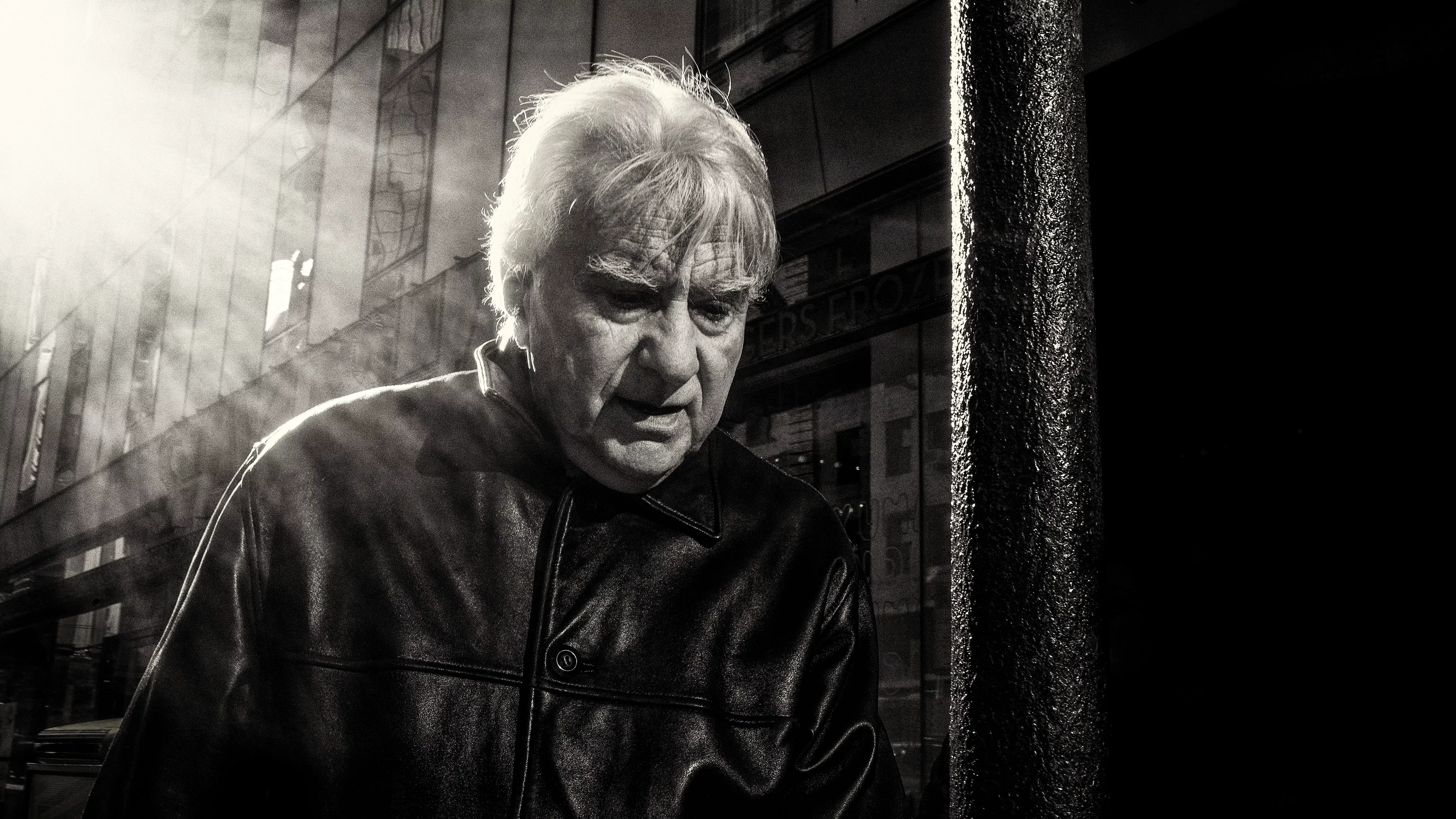 A black-and-white photograph of a man walking through the street. The sun shines over his shoulder.