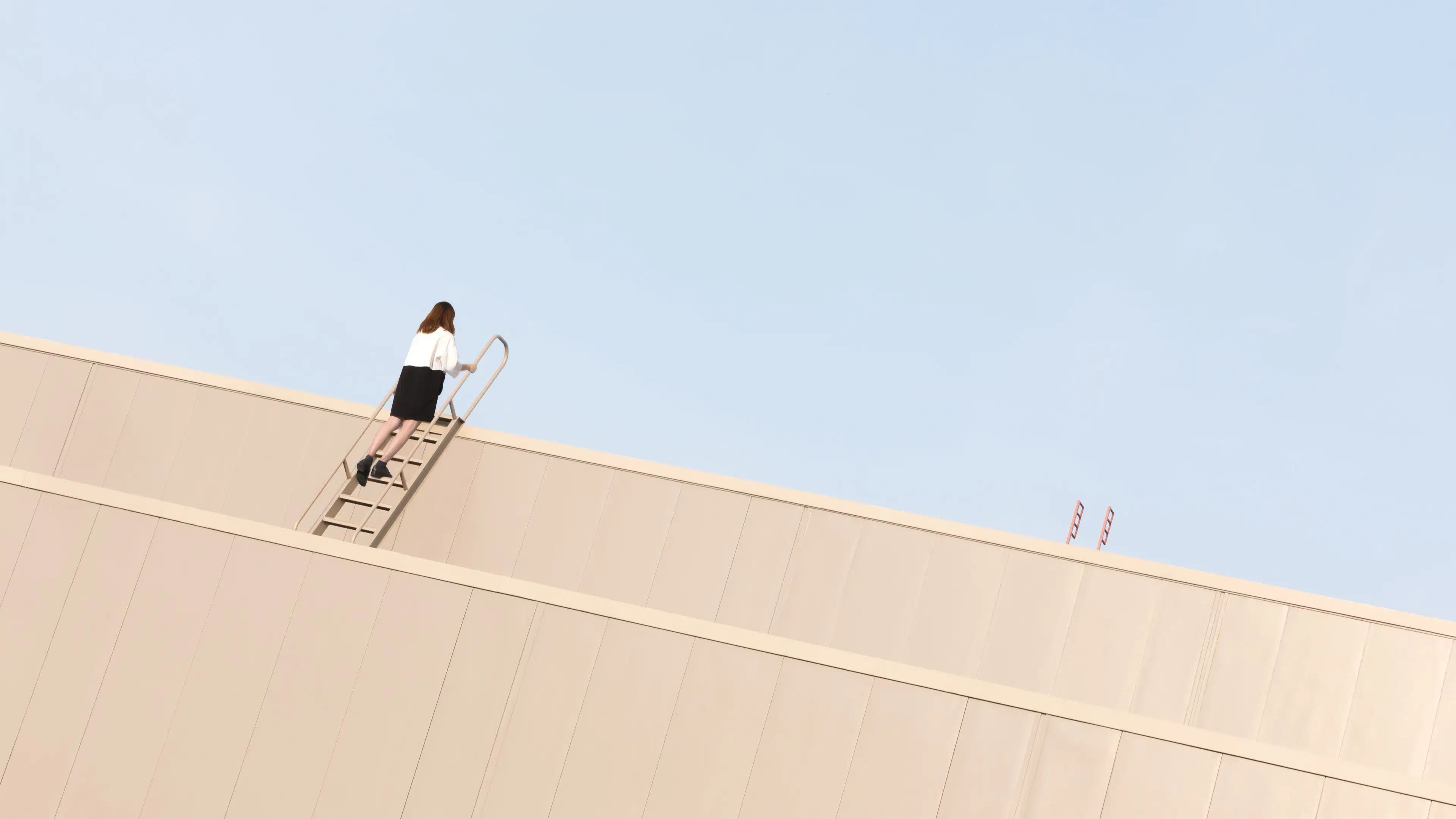 A photograph of a tan building jutting into a blue sky. There is a ladder coming down from the top of the building. Climbing up it is a woman with dark hair wearing a white shirt and black pants. 