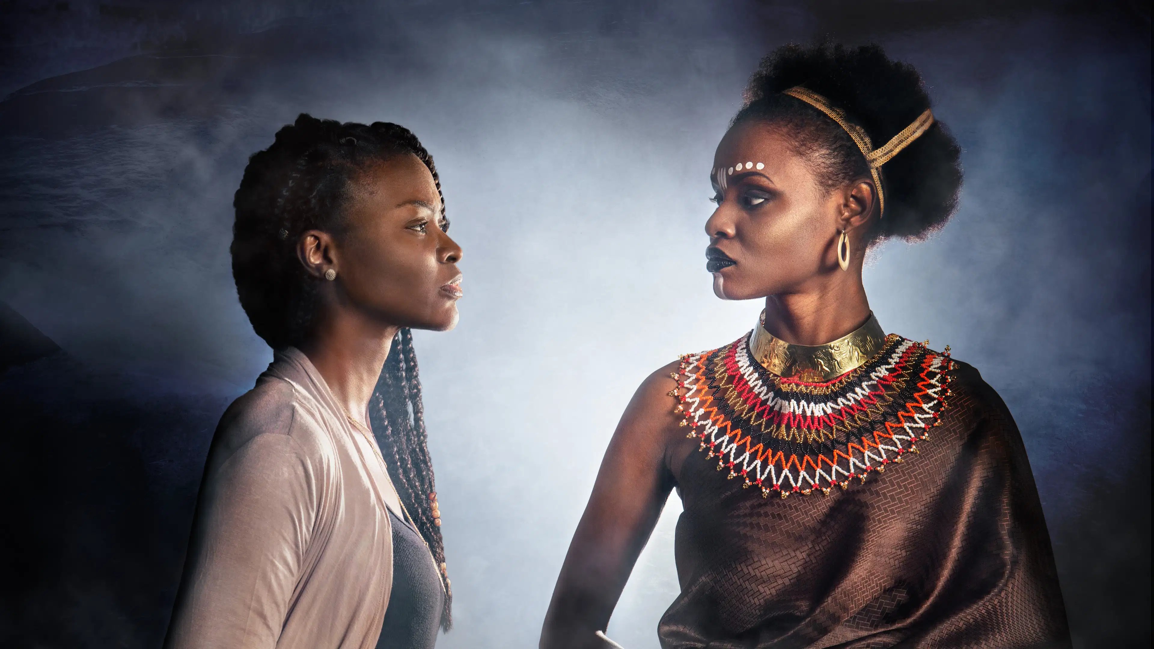 A portrait of two black women looking at each other. The woman on the left is wearing a sweater and has her hair in braids. The woman on the right is in traditional tribal attire and jewelry. The background is illuminated by a light behind them, and they are surrounded by fog.