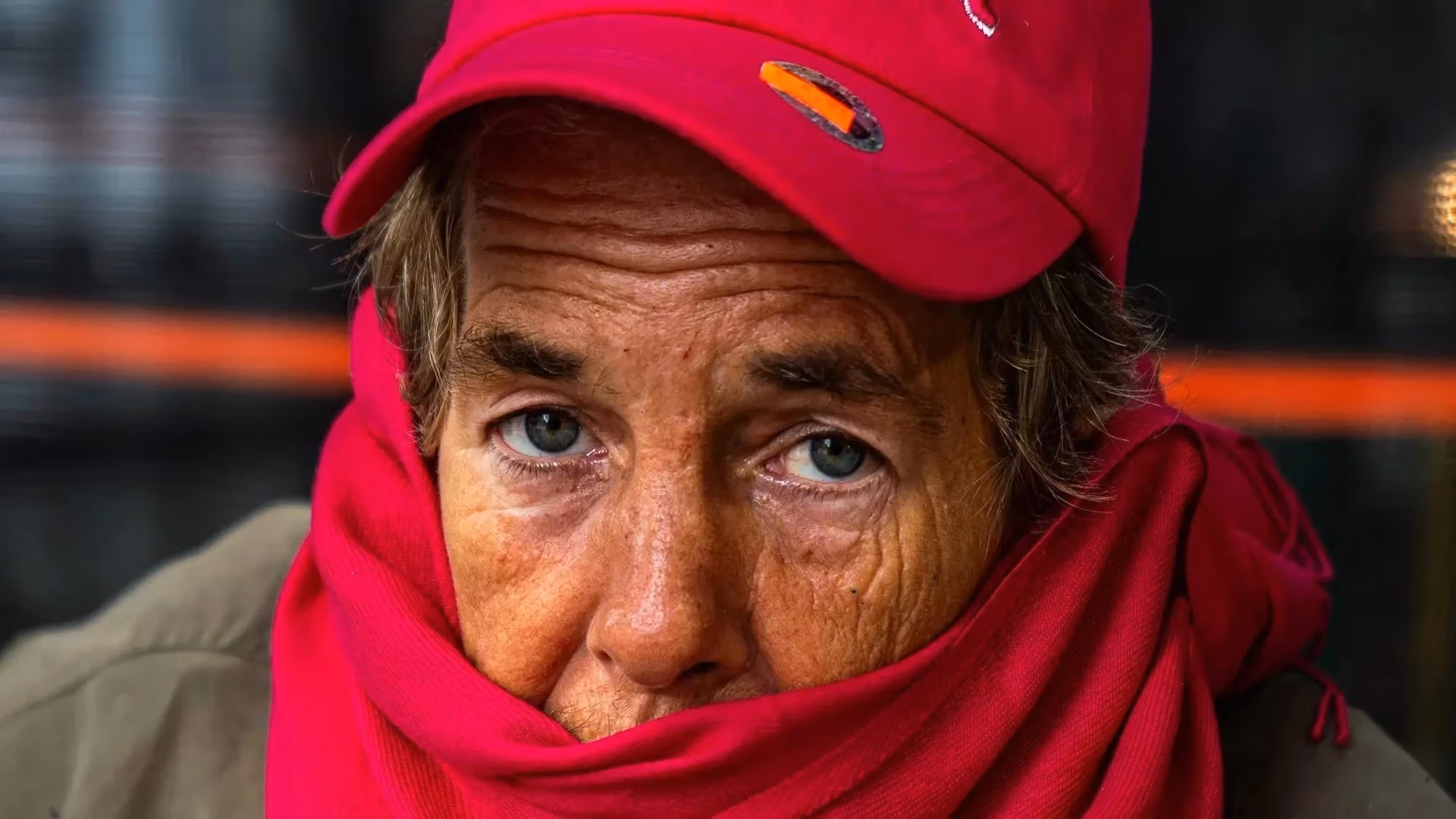 A portrait of an elder person with blue eyes and wrinkles on their face. They wear a red hat, red scarf and green coat.