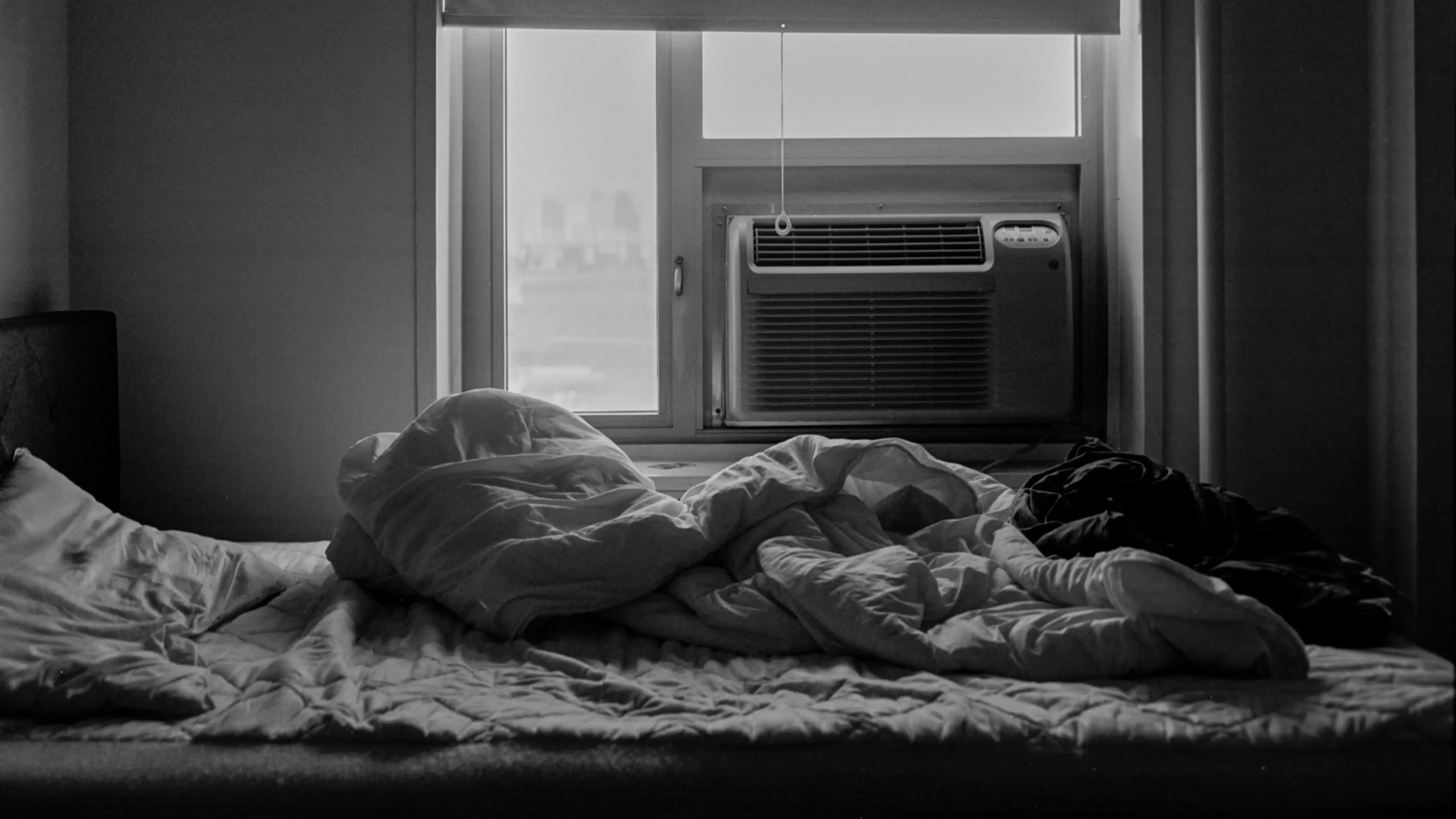 The black and white photo shows an unmade bed and a window air conditioner under half-drawn curtains
