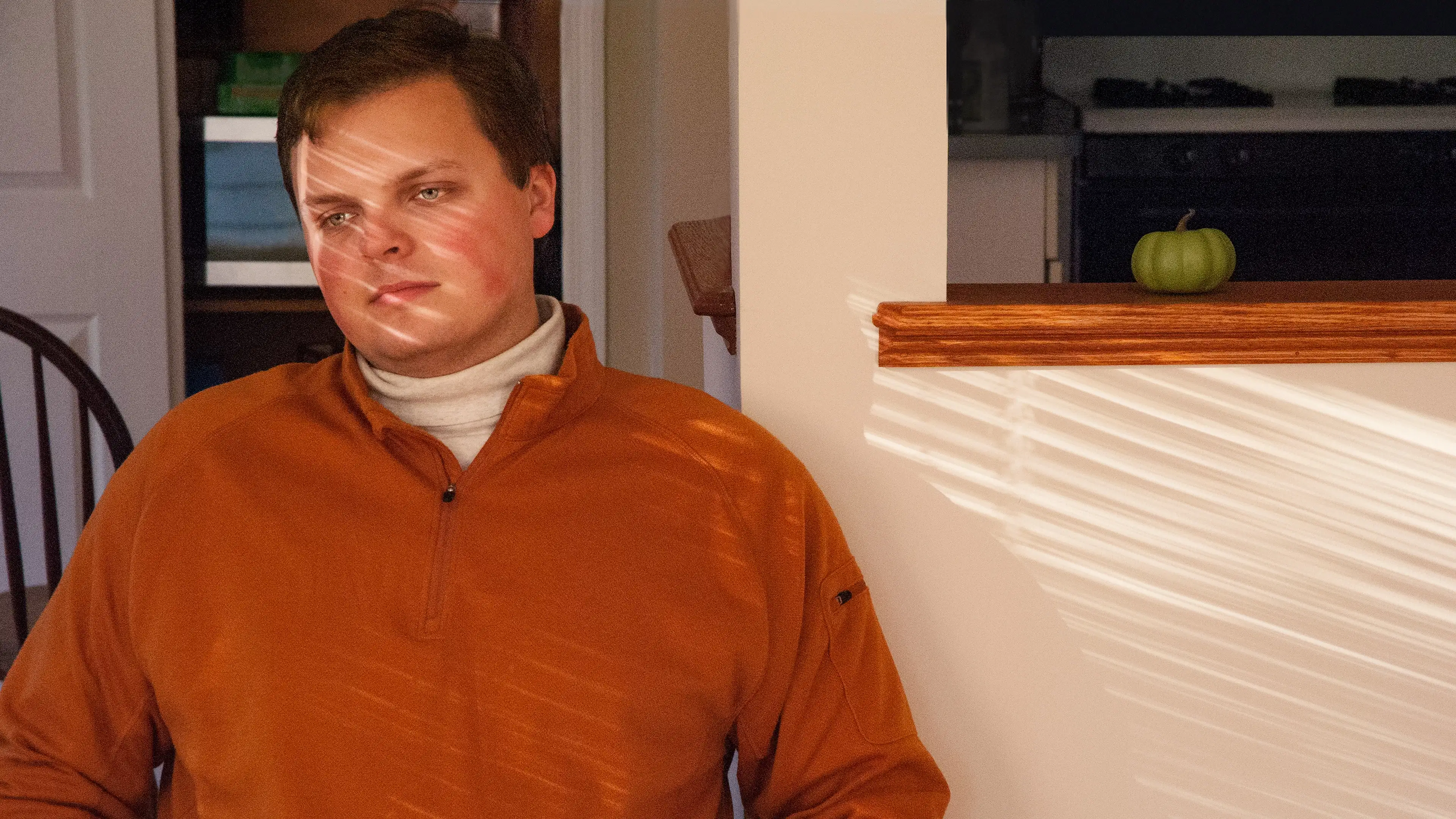 A photograph of a man from the waist up wearing an orange sweater, staring off the left frame of the camera. He is sitting in a house, with a wall and hair behind him. There is a reflection of sunlight from blinds streaking across his shirt and face and the wall next to him. 