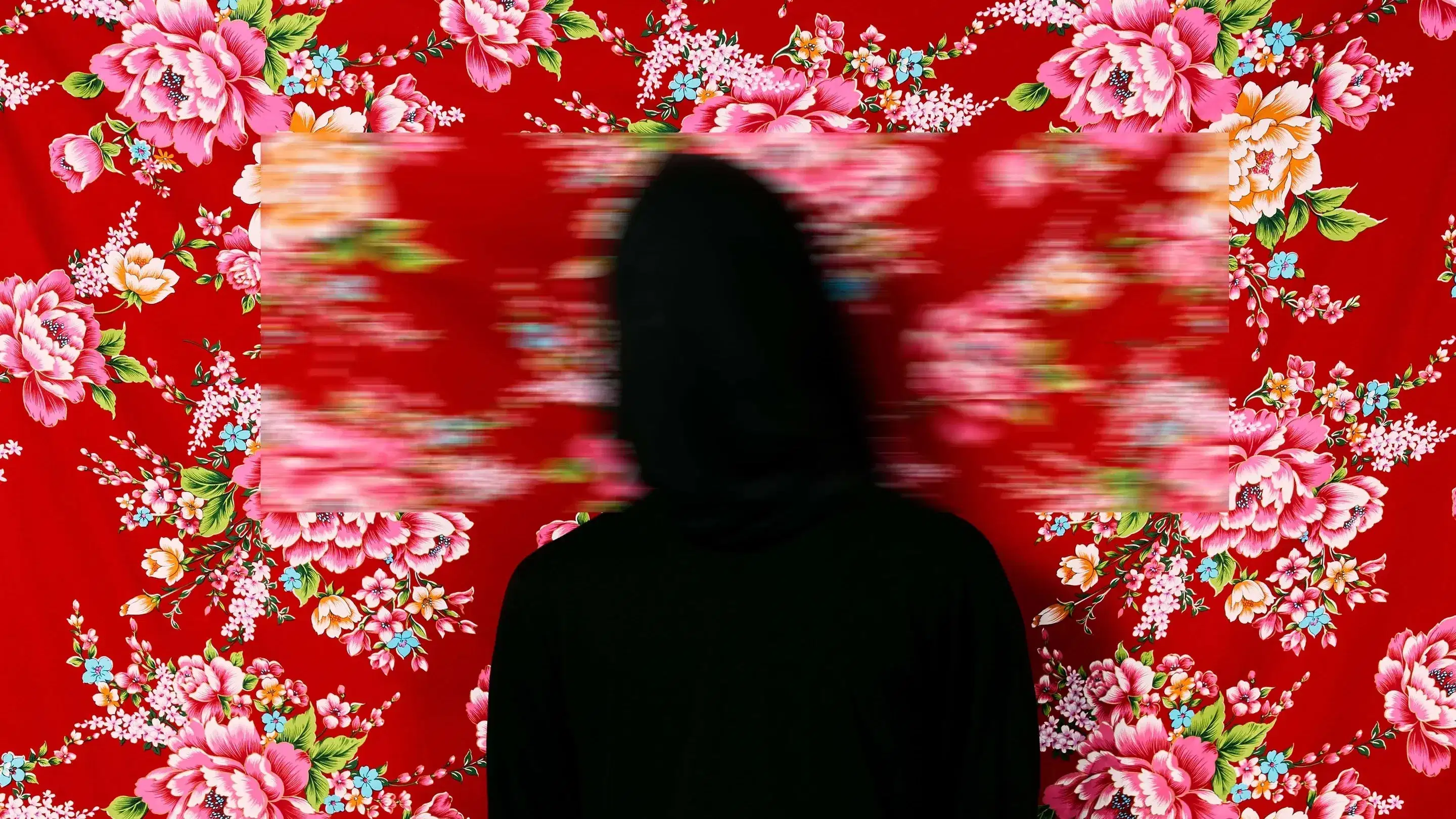 A color photograph featuring a figure wrapped in black from head to toe standing facing the camera against a red Hakka floral fabric background, which is suspended and reveals a faint dark wooden floor below. Three blurred rectangles cover the head, hands, and feet.