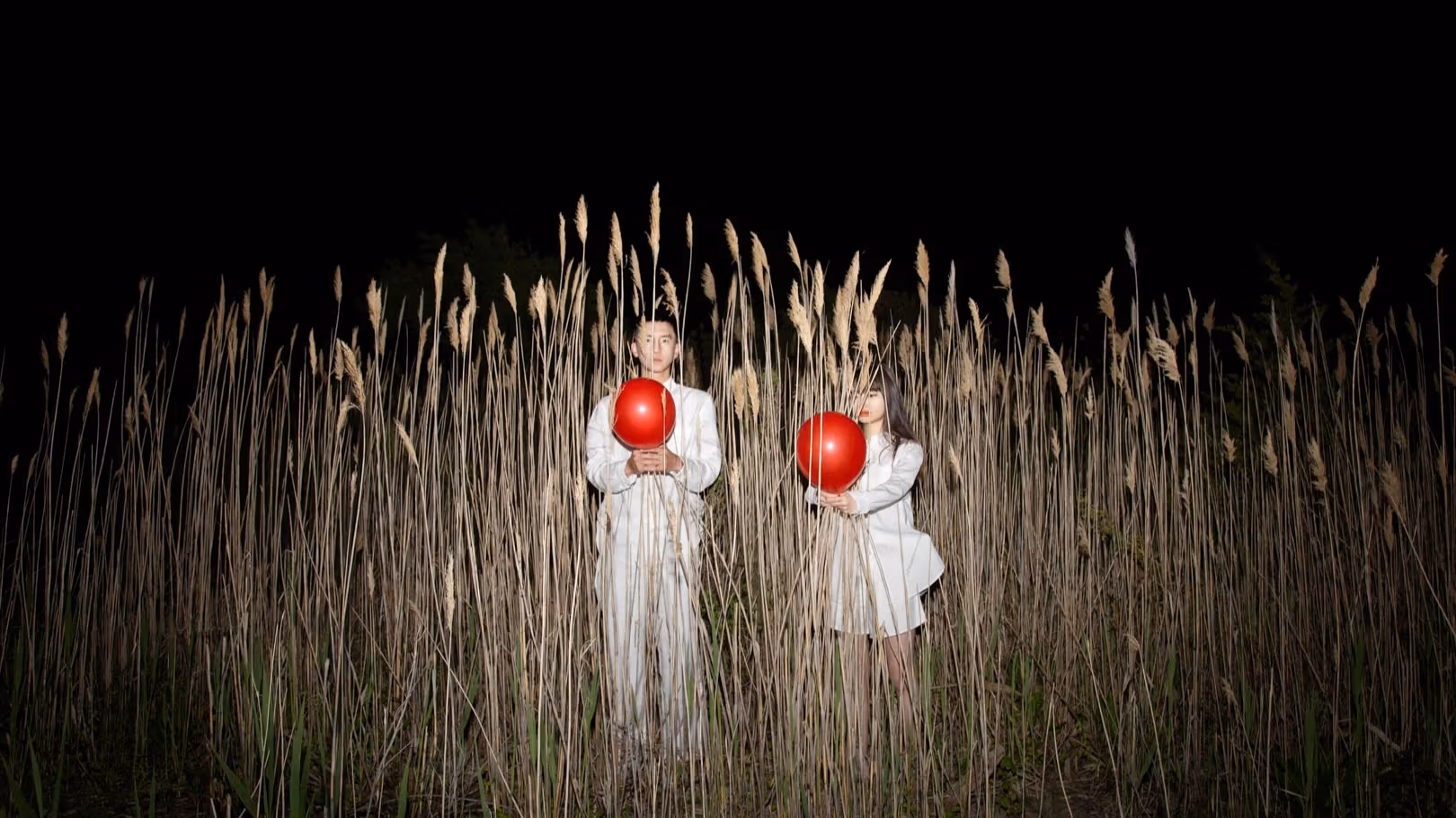 A man and a woman, both wearing white clothes and each holding a red balloon, stand in the middle of tall reeds at night.