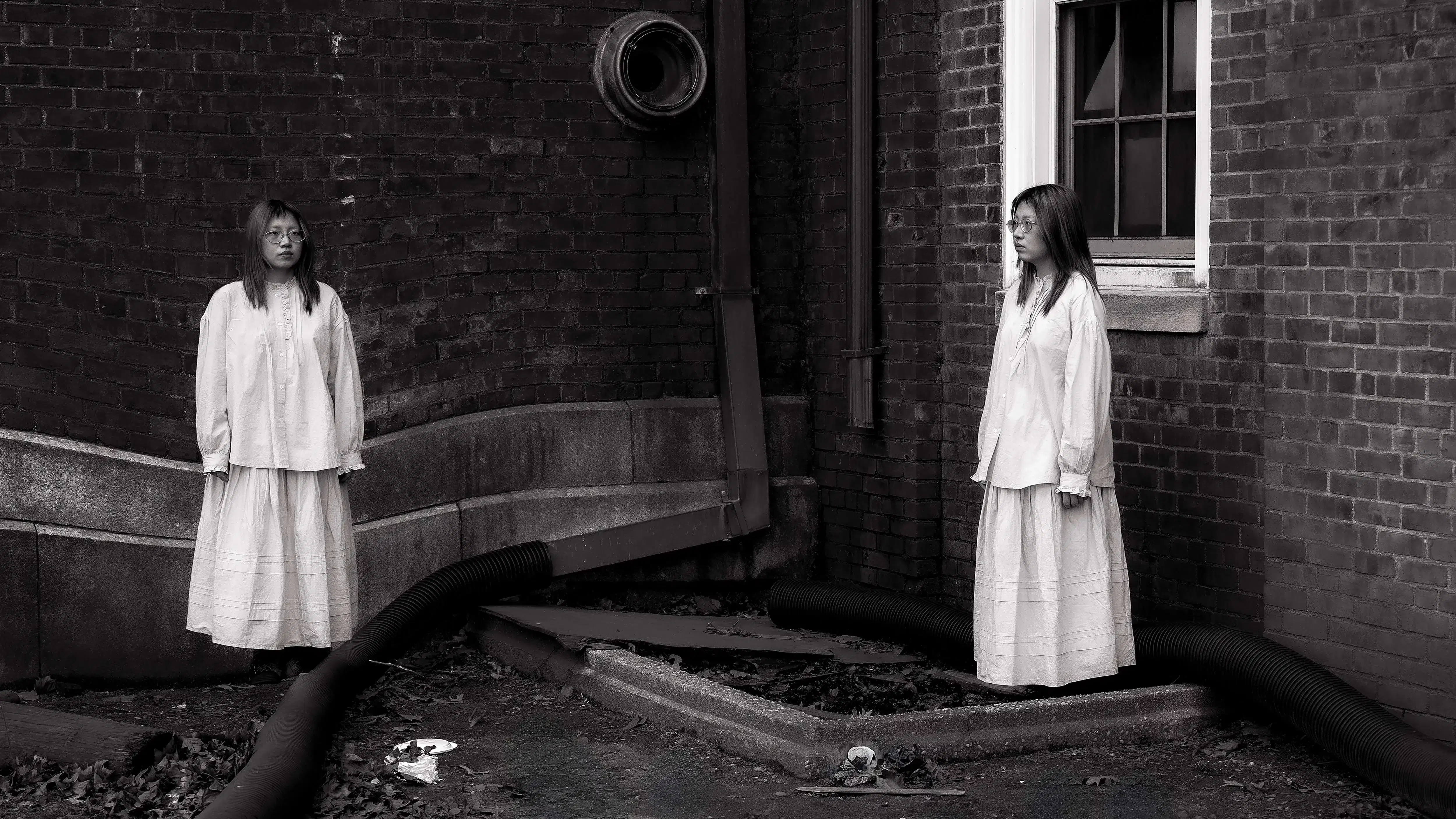 Black and white photograph of a girl duplicated in the frame, standing at two corners of a brick building. Both wear the same long dress, facing each other across a barren, debris-littered ground with industrial pipes.