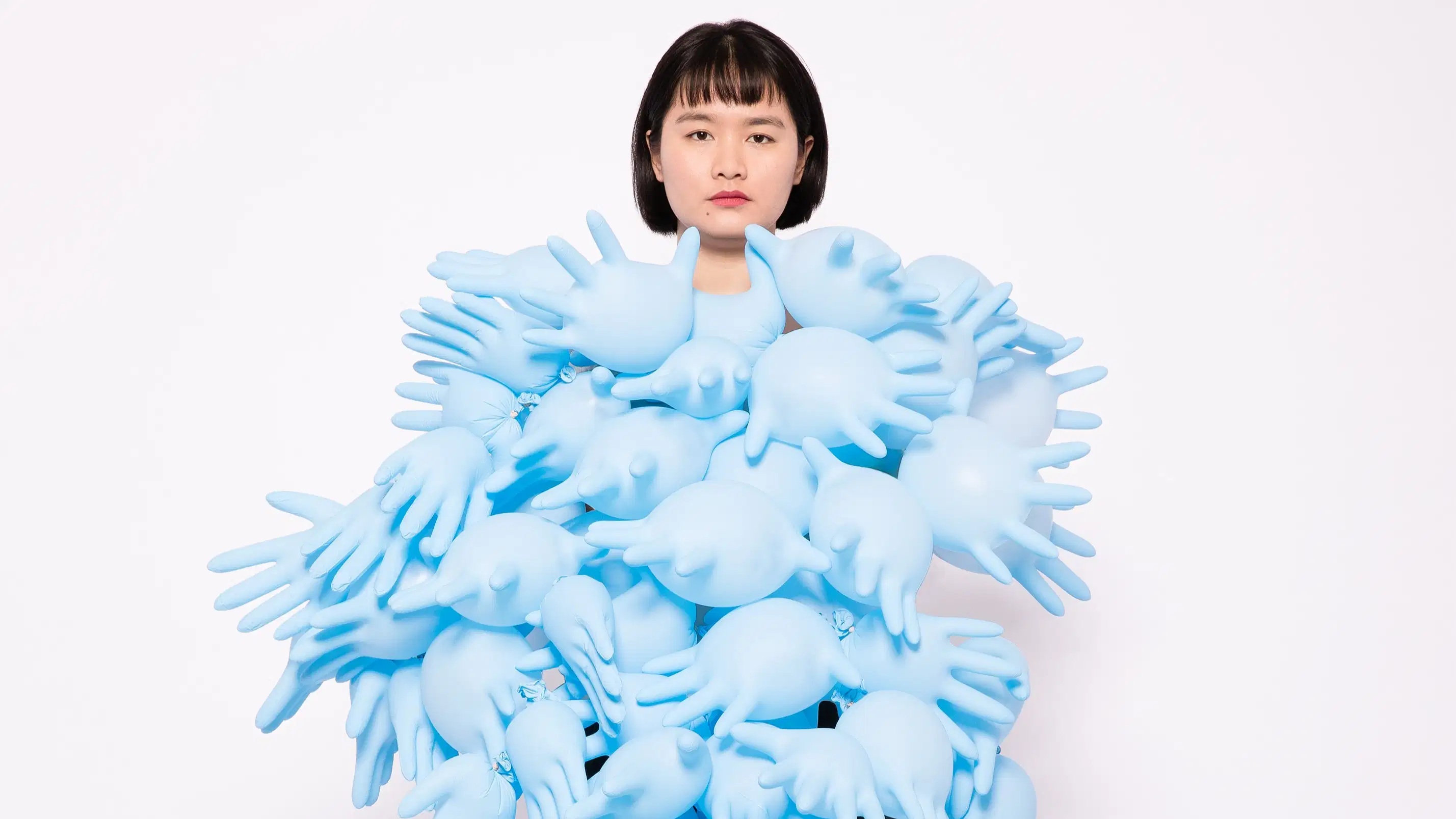 A studio photograph of a woman standing against a white background. She is covered in blue, blown up latex gloves from neck to feet.
