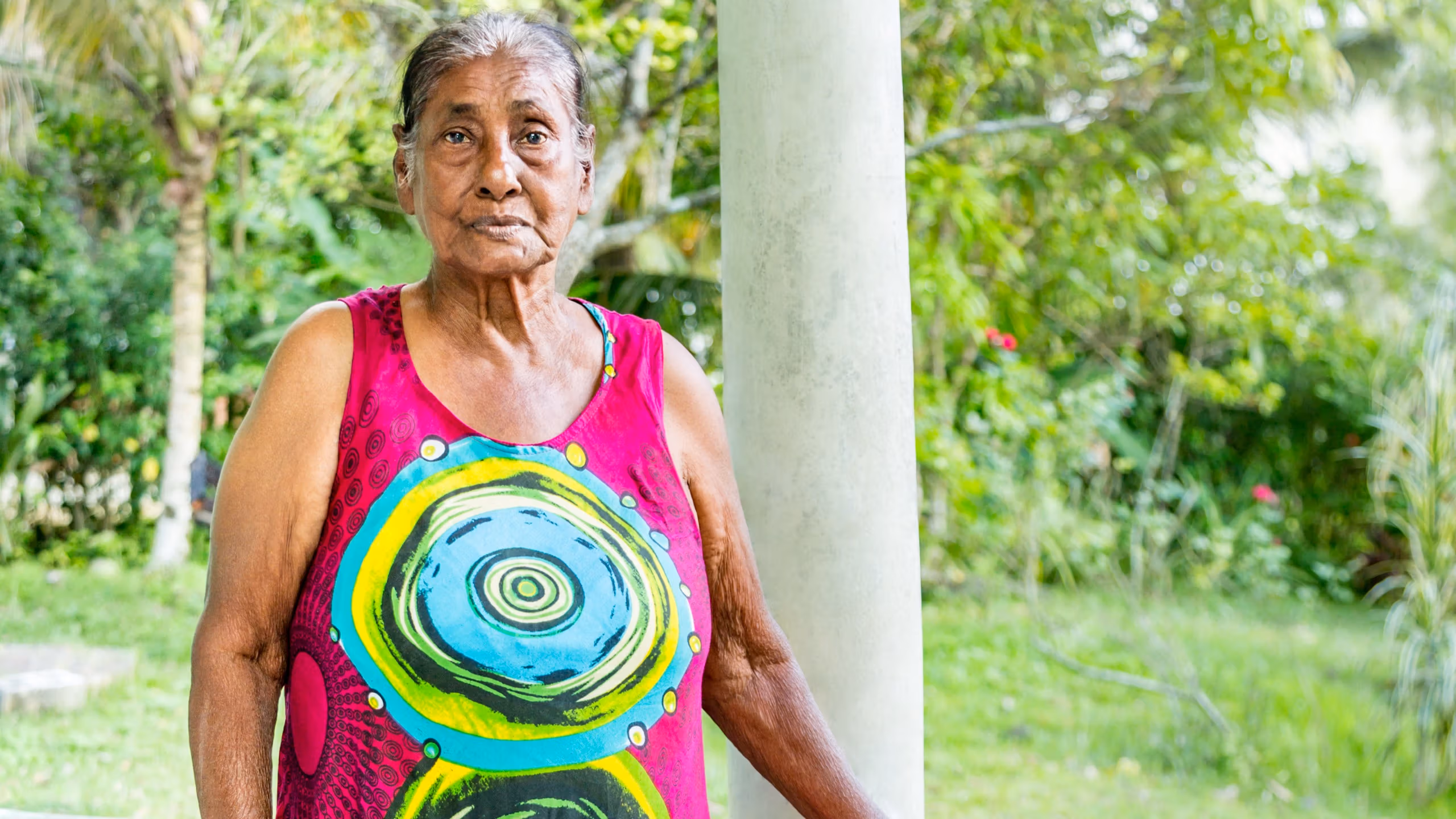 A photograph of an older woman standing with her hand resting against a teal colored wall. There is a white column behind her. She is standing under a structure outside. She is wearing a pink dress with colorful circle designs on it and blue shoes. She is staring at the camera.