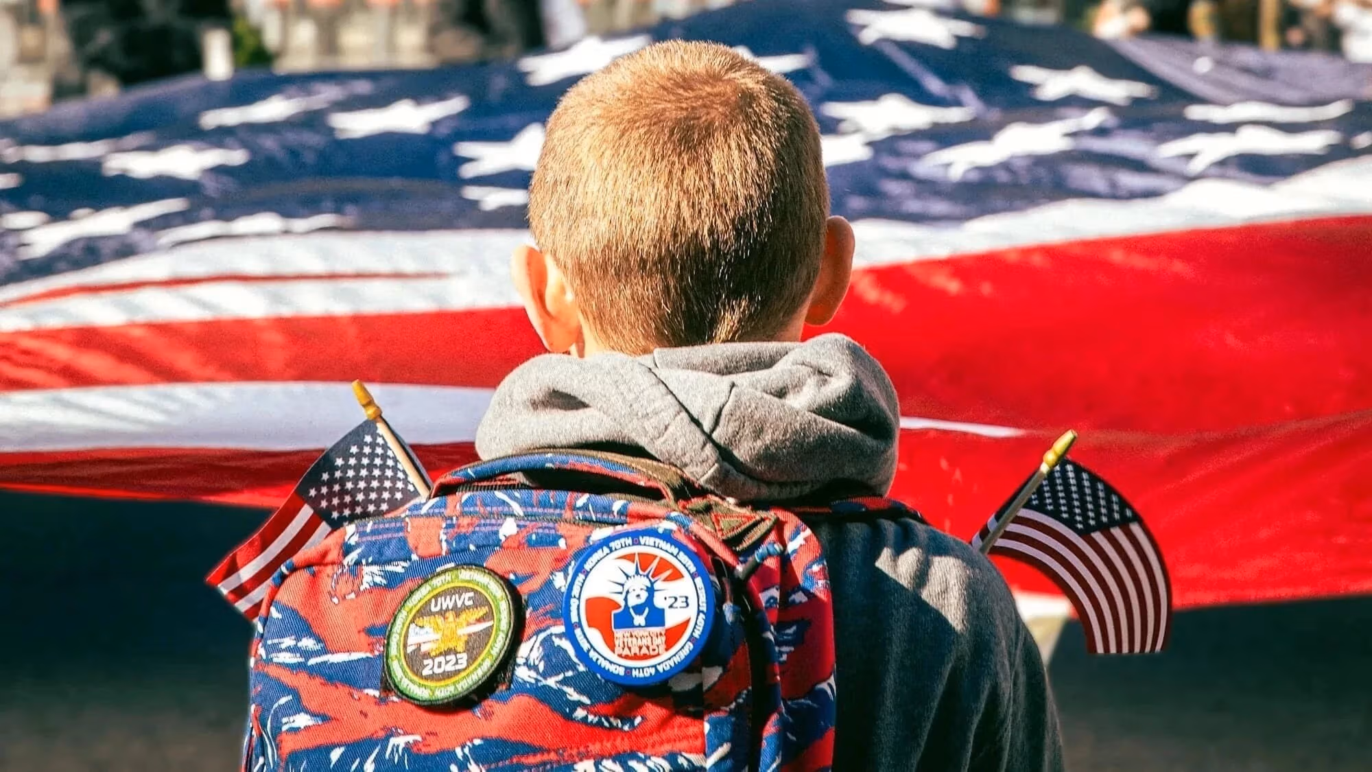 A kid faces away from the camera to look at a large american flag. He has patches on his backpack and holds two small american flags.