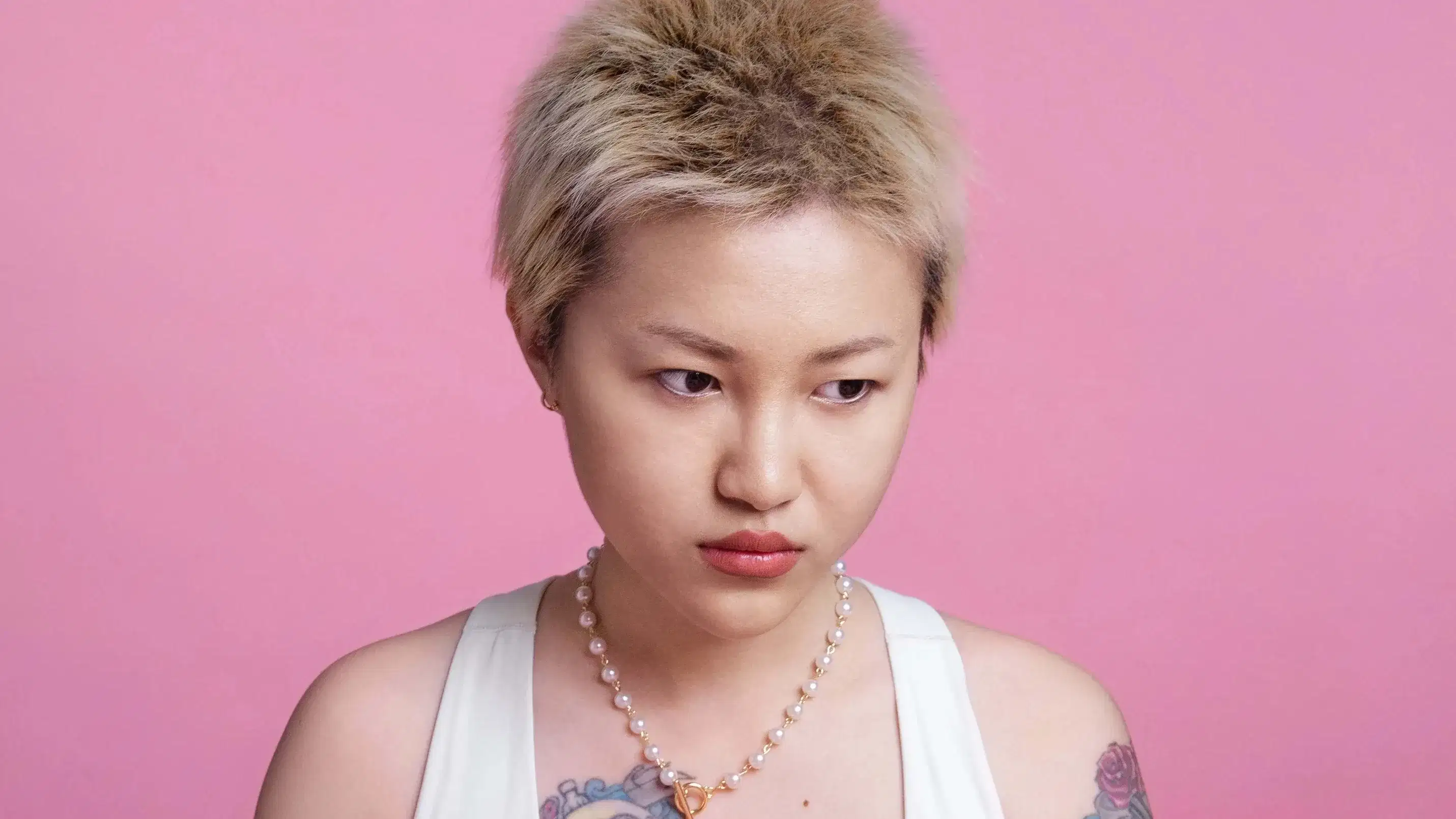 In this color photograph: On a pink background, a half-length portrait of a gold-haired young Asian woman wearing a white sports bra, sitting. She has large colorful tattoos on her right chest and left arm. On the pink table in front of her are three red  tomatoes. She is holding a smaller dark- colored tomato and is looking away  from the camera, appearing thoughtful.