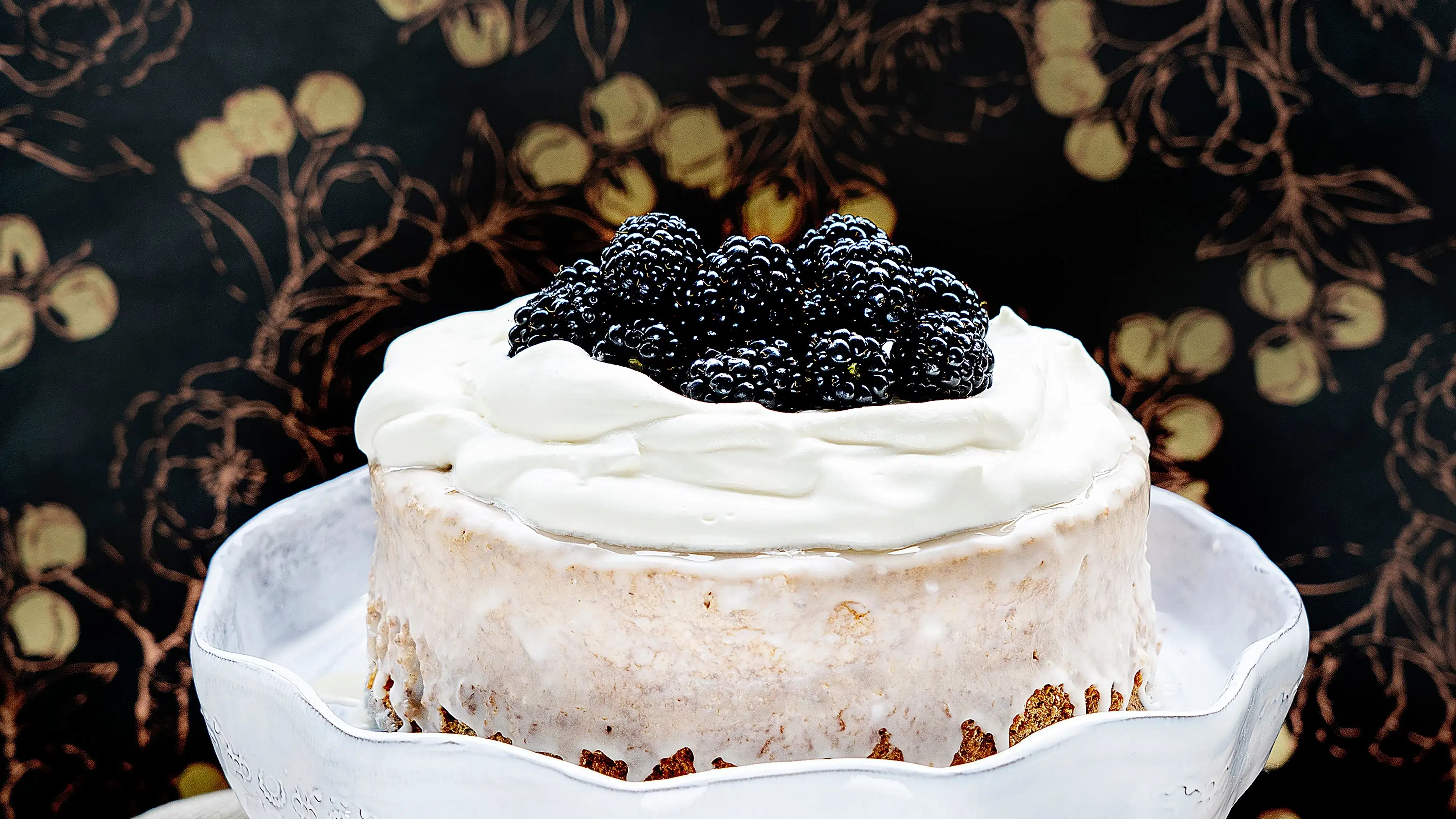 A cake that is covered with sugar glaze and topped with whipped cream and blackberries on a white cake stand. The background it dark with a gold pattern and there are Christmas ornaments and a silver fork next to the cake stand.