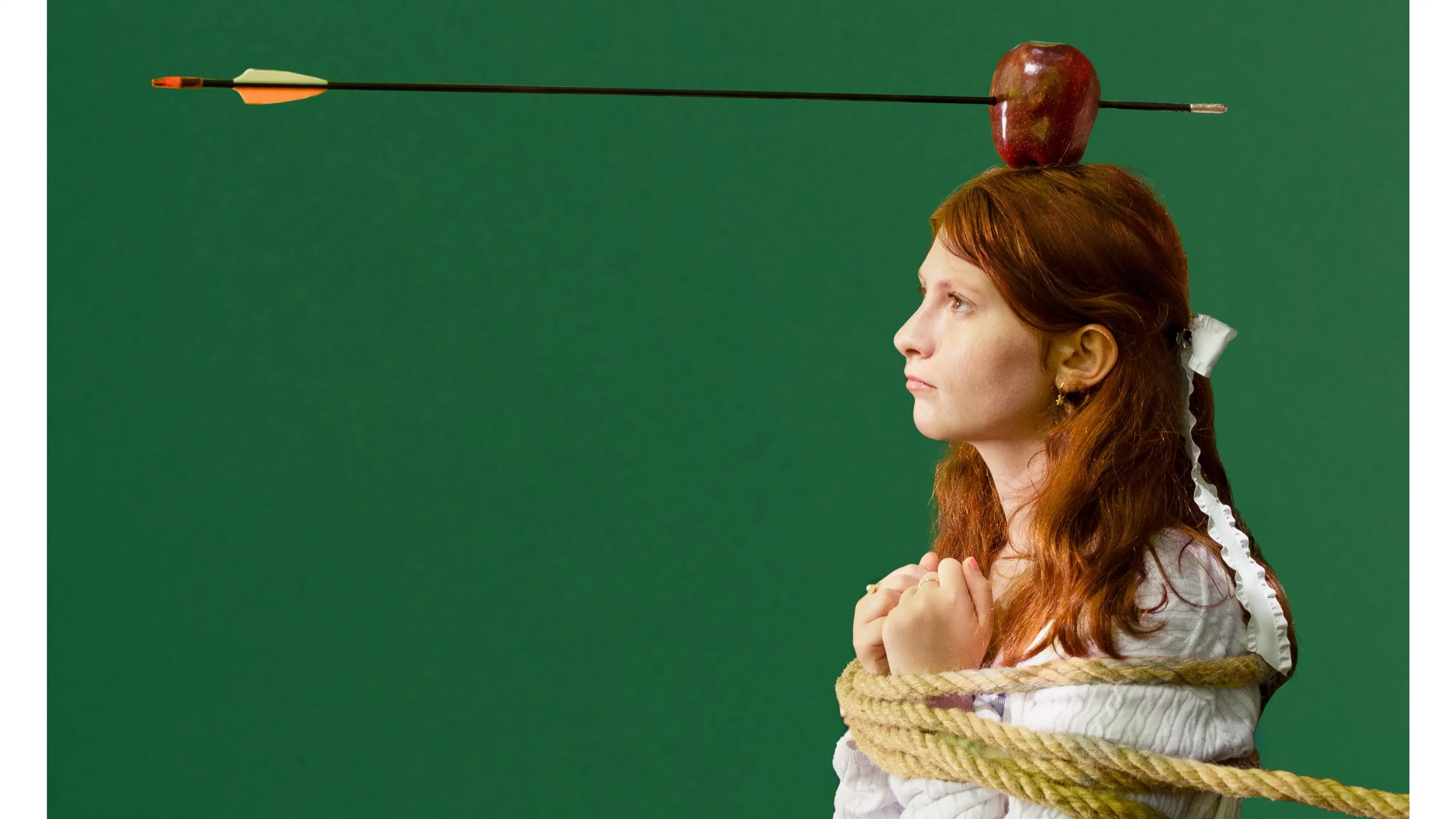 A teen girl in profile is bound by a rope. There is an apple on her head with an arrow shot partway through it.