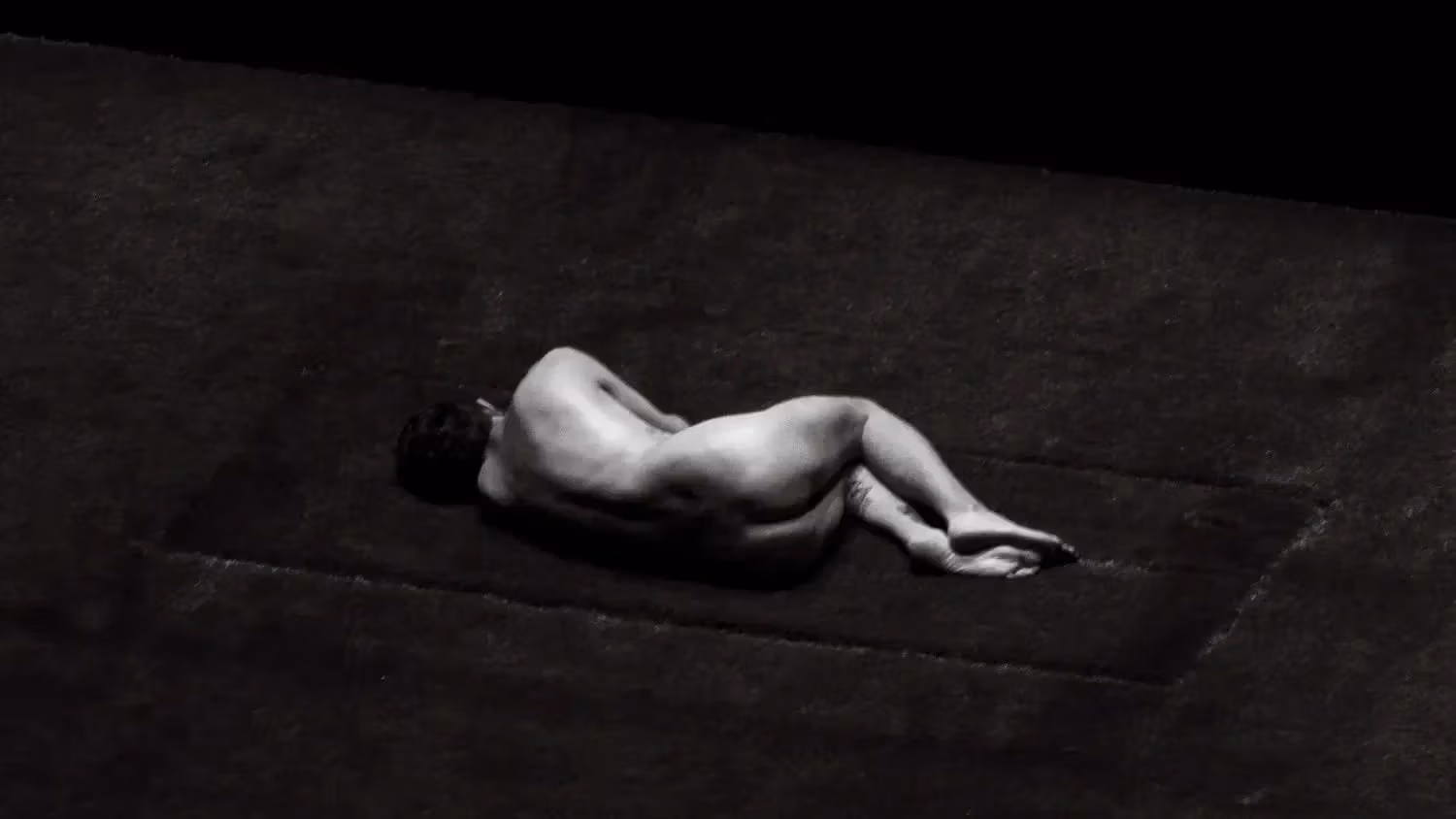 black and white image of naked man from behind in curled position, laying on grass. 
