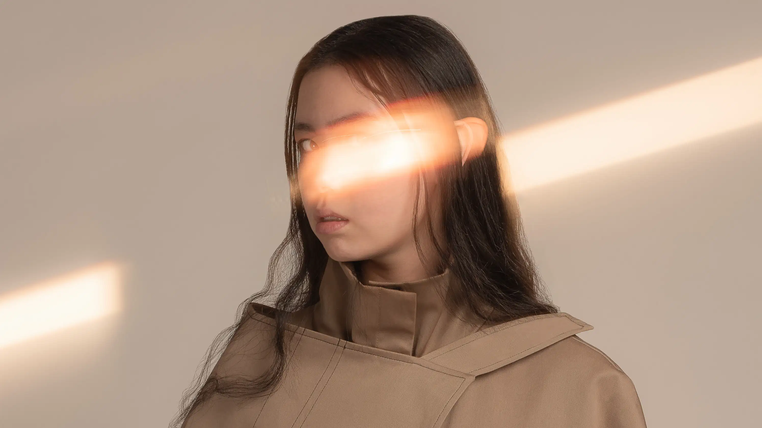 A photograph of a woman with dark brown hair in front of a tan background. She is wearing a tan jacket covered in buckles and pockets. SHe is looking at the camera. There is a streak of light going across the frame and over the model’s face, covering her nose and left eye.