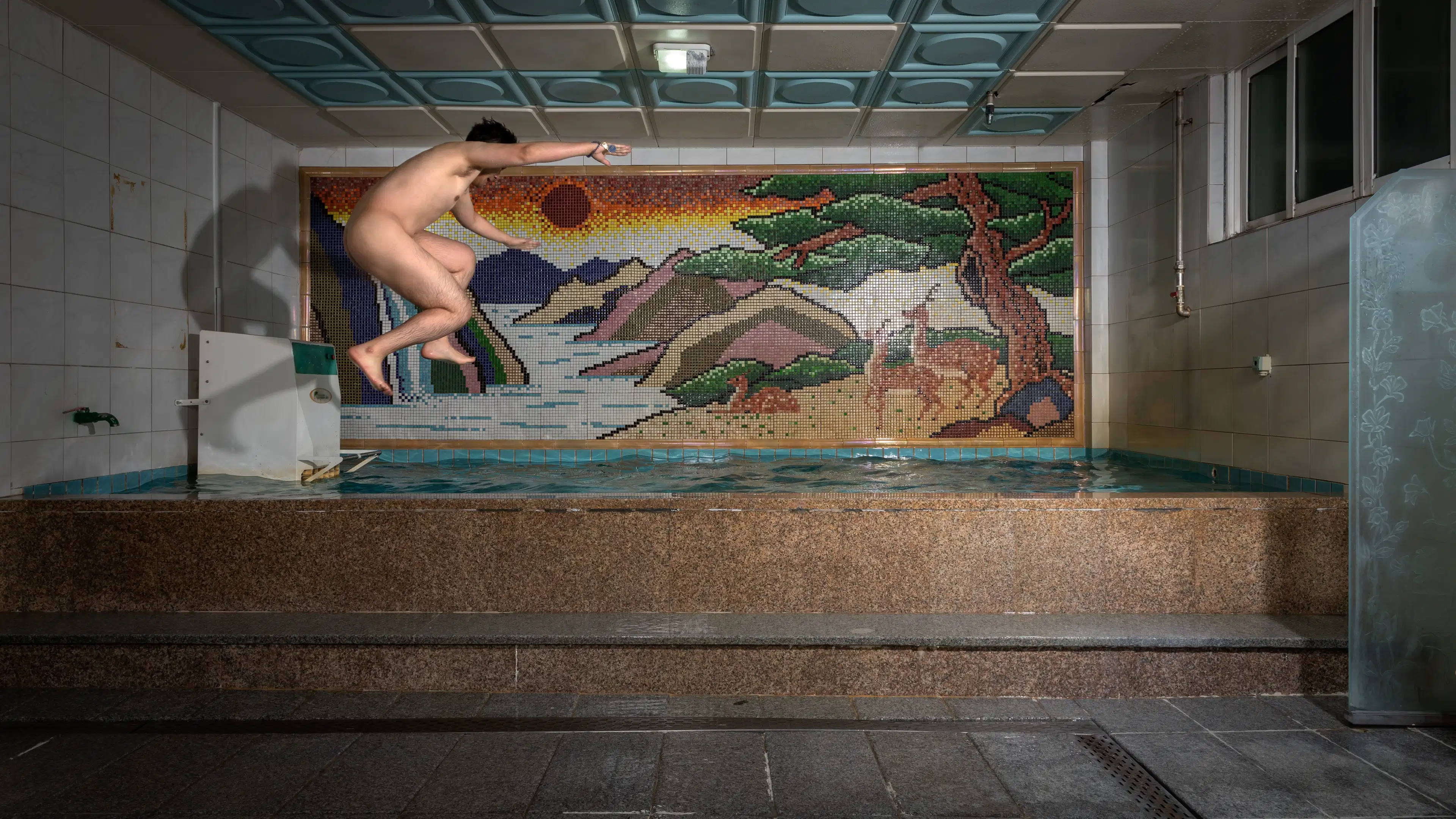 A documentary photograph of a nude man in profile, jumping from left to right side of a large tiled bath into the water. On the wall behind him is a colorful tile mosaic of a landscape.