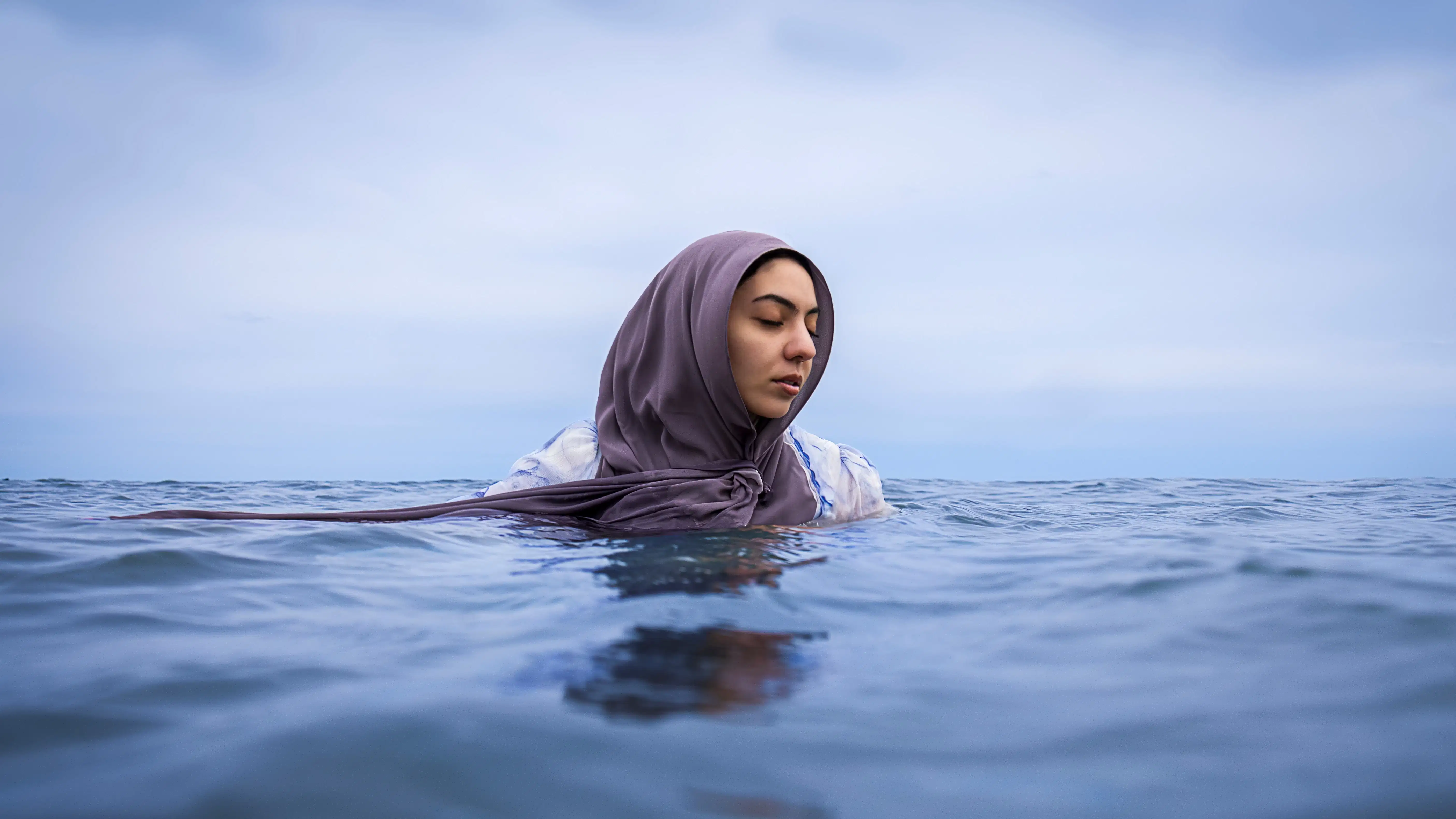 A woman wears a light purple hijab and is partially submerged in calm ocean waters. The horizon line divides the frame, with the vast, tranquil sky above and the gently rippling water below. The woman has a serene and contemplative expression, her eyes closed as she floats.