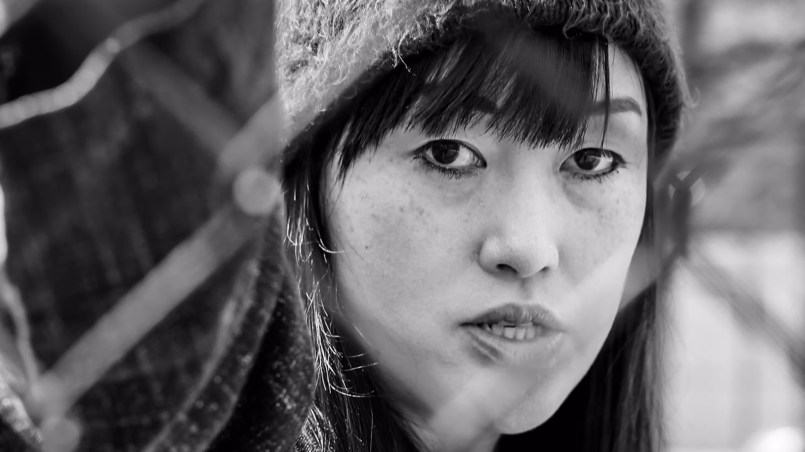 A black and white photograph of a woman looking at the camera through a chainlink fence. She has her left hand up against the fence, off frame. She is looking at the camera with her mouth slightly parted. She has dark, straight hair with bangs, and is wearing a beanie, a dark unzipped sweatshirt, and a white shirt. 
