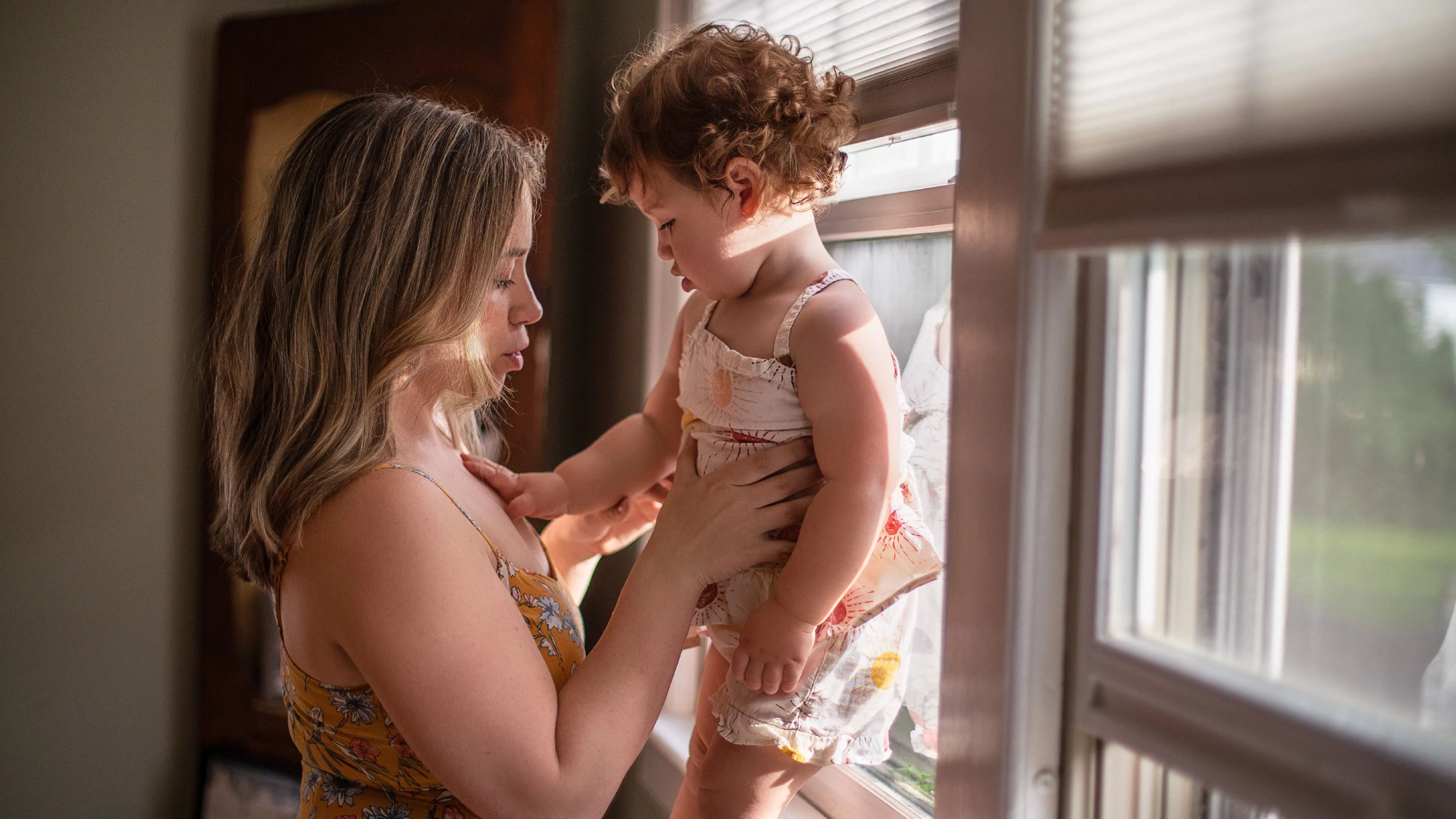 A woman holding her baby who is standing up next to the window, the baby’s hand in touching the woman’s chest. They both are looking at the baby’s hand. The woman is wearing a floral dress and the baby a floral jumpsuit. Her legs are not visible. 