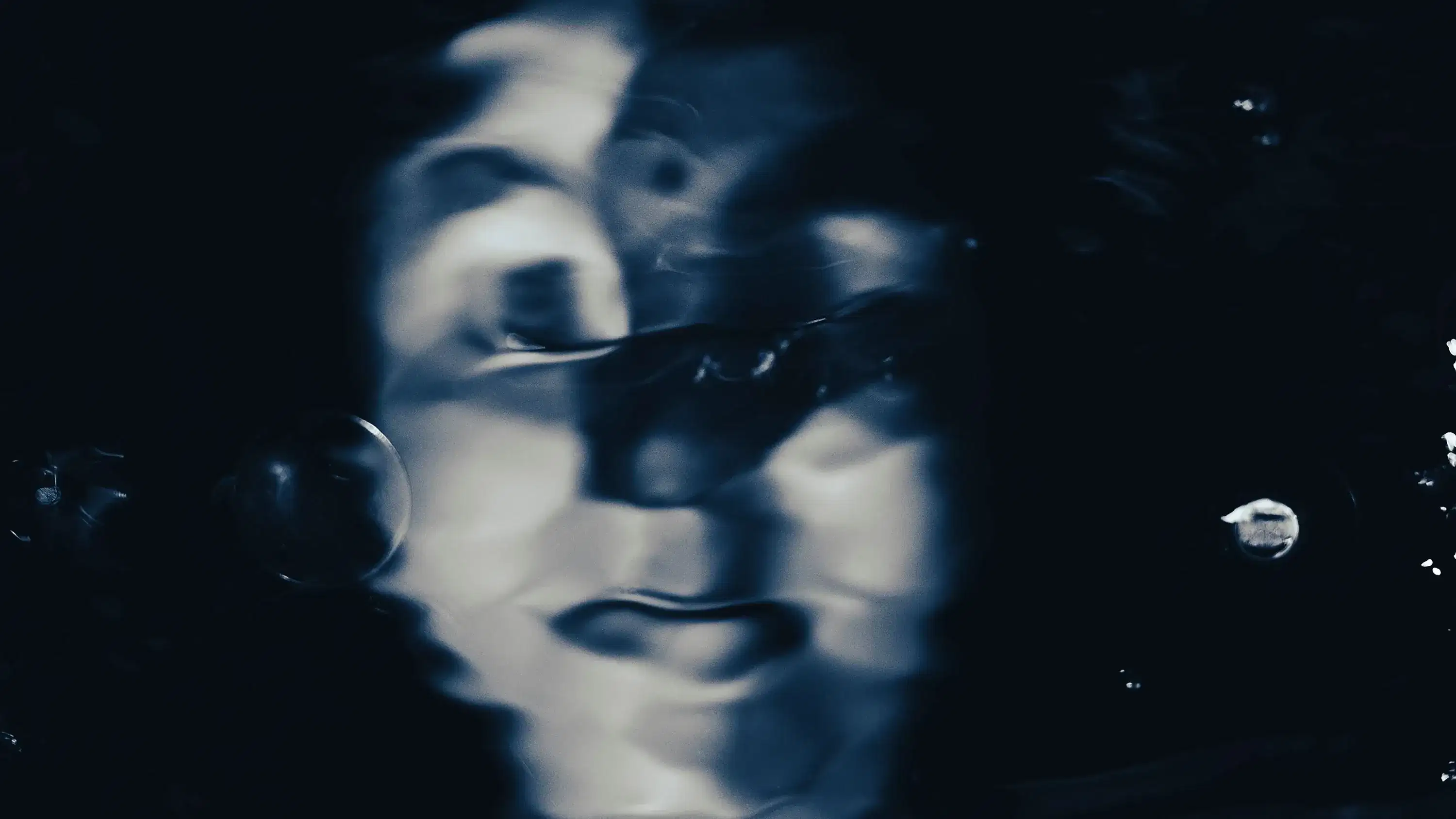 A blurred female face with eyes half closed and a bindi in between her eyebrows appears submerged from a distance, with bubbles and reflections creating a soft distortion. The image has dark navy and pale blue hues.