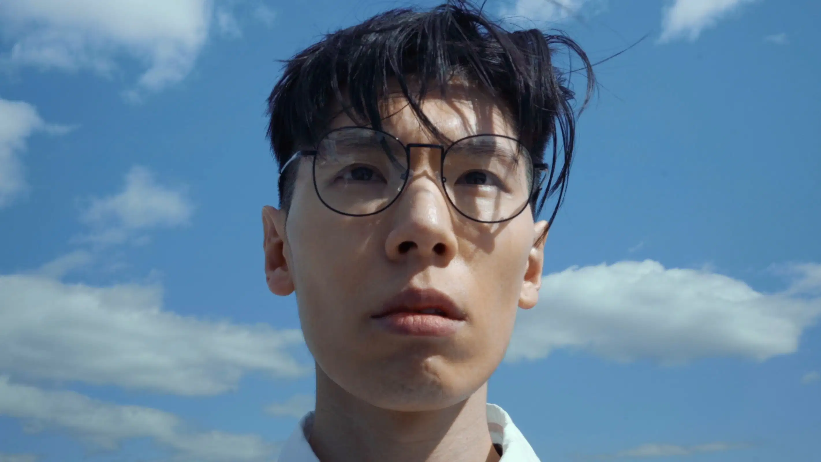 A close-up of a man's face looking off-camera. Behind him is a blue sky with clouds.