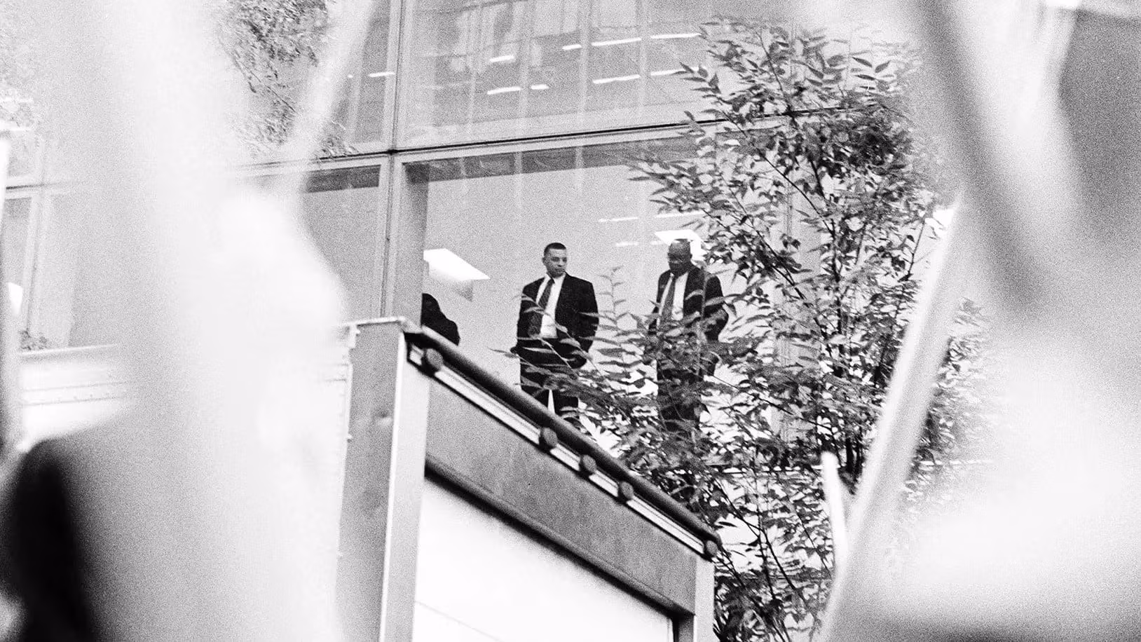 Two men in suits observe a protest from an elevated position