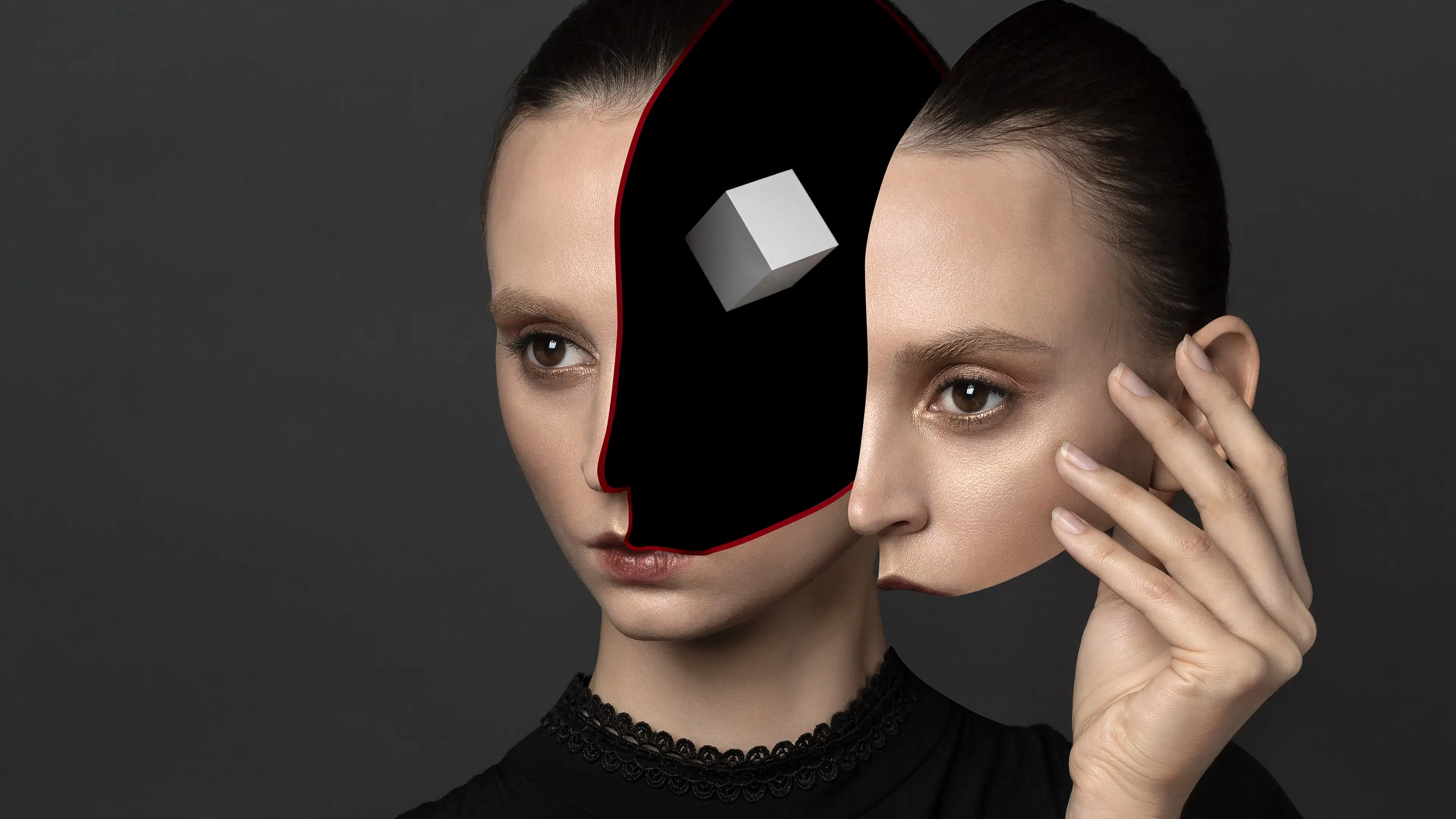 A studio headshot portrait of a woman against a gray background, wearing a black blouse. The left portion of her face from forehead to mouth has been removed, leaving a black negative void. Suspended in the void is a small, white cube. The woman’s right hand is held up next to her head, and holding the removed portion of her face like a mask.