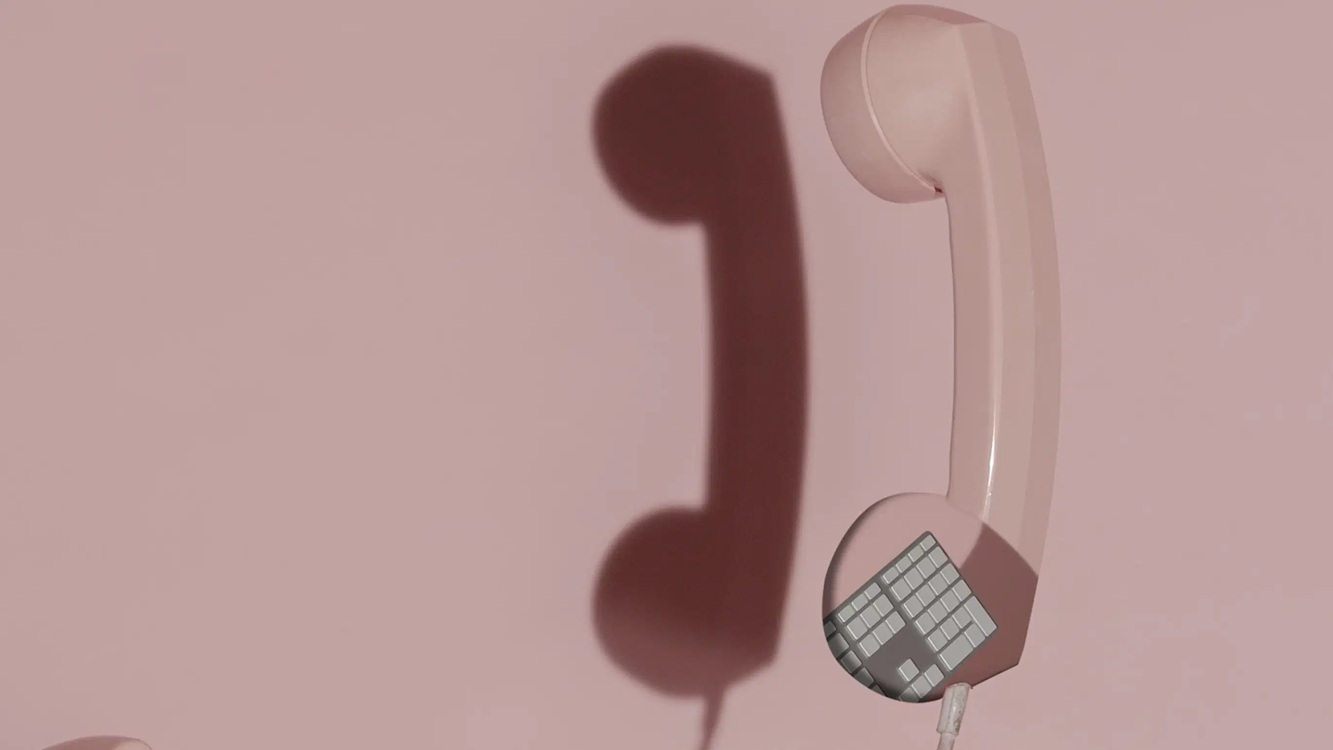 A muted color photograph of a pink rotary phone suspended in front of a pink background.