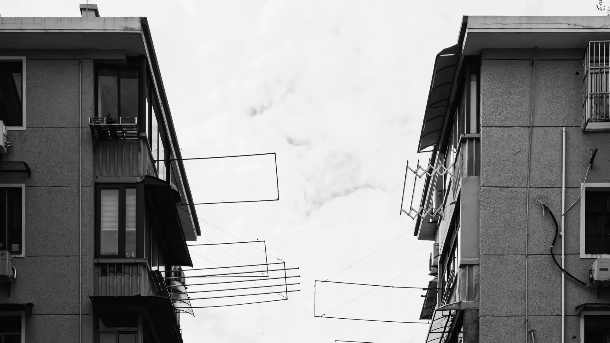 A black and white photograph capturing a narrow gap between two residential buildings. Multiple rows of empty clotheslines extend from the balconies, creating a pattern of horizontal lines against the sky. 
