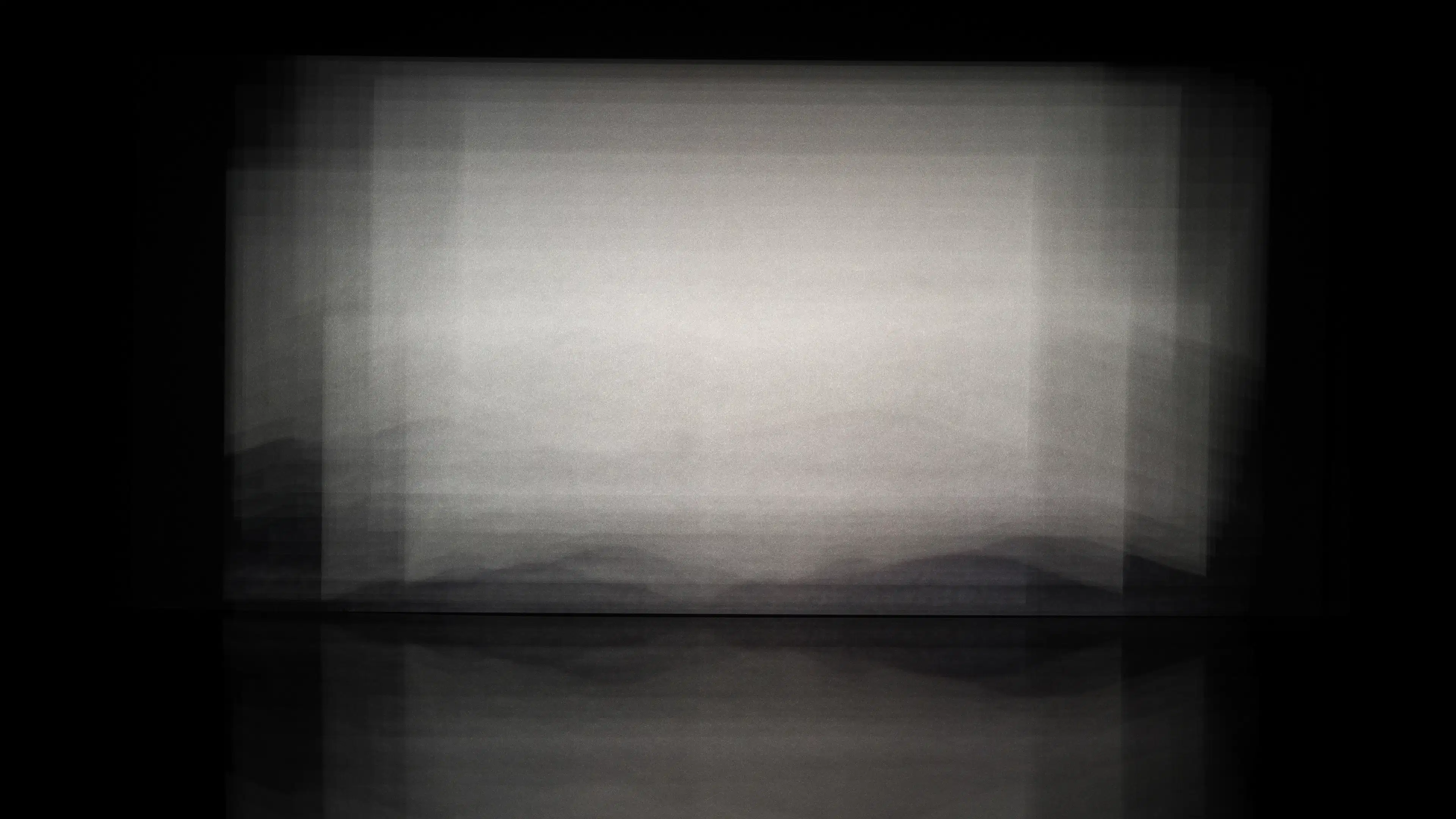 An abstract photograph of a square surrounded by a black background. The square is grey, and it is blurred and foggy, with no discernable shape to it. There are faint images of hills within the square.