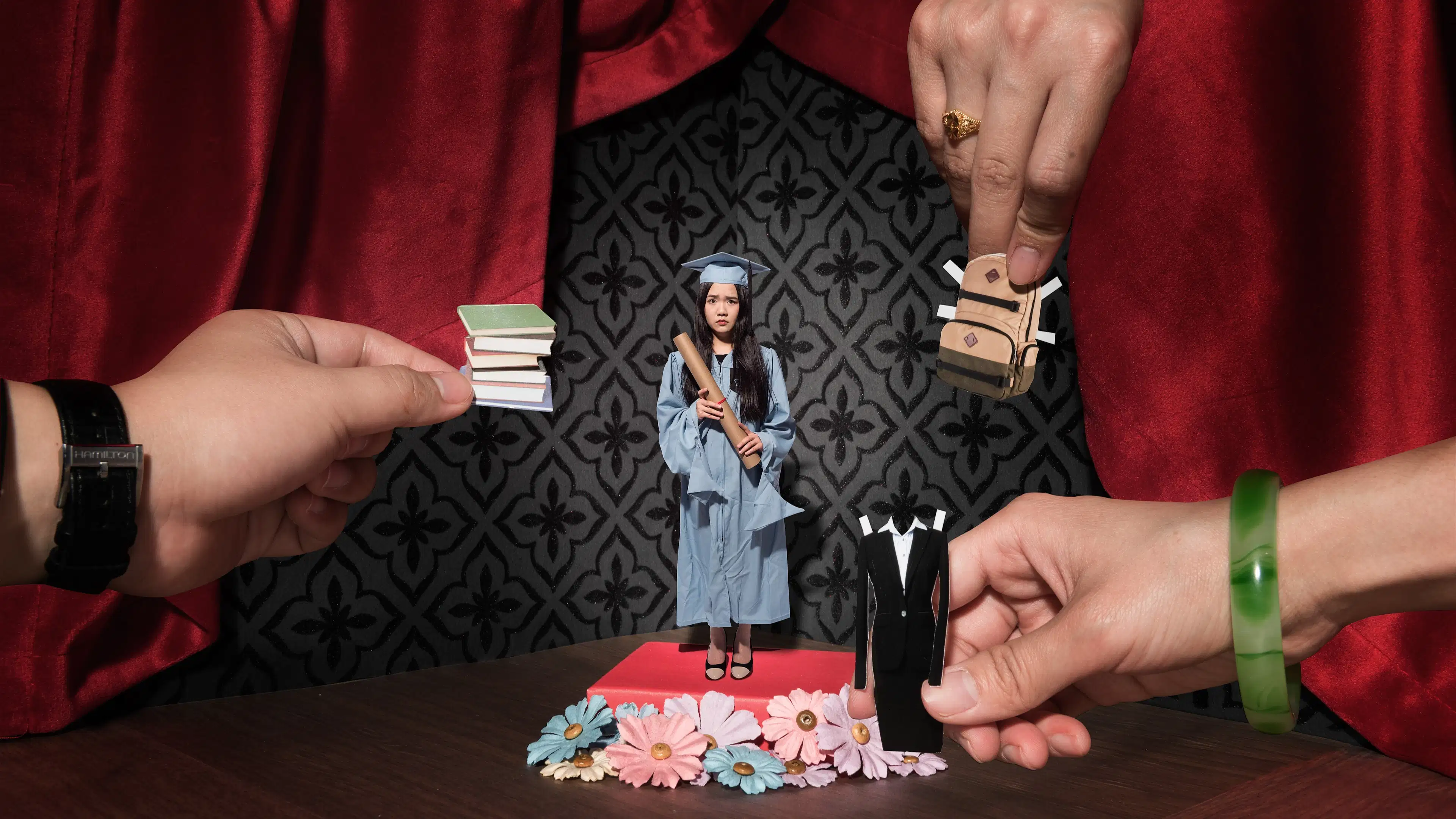 A photograph of a diorama of a room. It has a wood floor, grey wallpapered wall, and red curtain around it. In the middle is a pink pedestal with flowers around it. On the pedestal is a woman in a blue graduation gown and hat, holding a diploma. She is looking at the camera with a worried expression. There are three hands coming into the frame from different sides. One is holding paper cut out of a stack of books, one is holding a paper cut out of a backpack, and the last is holding a paper cut out of a business dress.