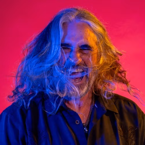 A portrait of Michael Kaminski in front of a hot pink backdrop. Michael has long, silver hair, which in the picture appears to be spun around. His face, along with his hair, is lit by a blue light on the left and an orange light on the right. 