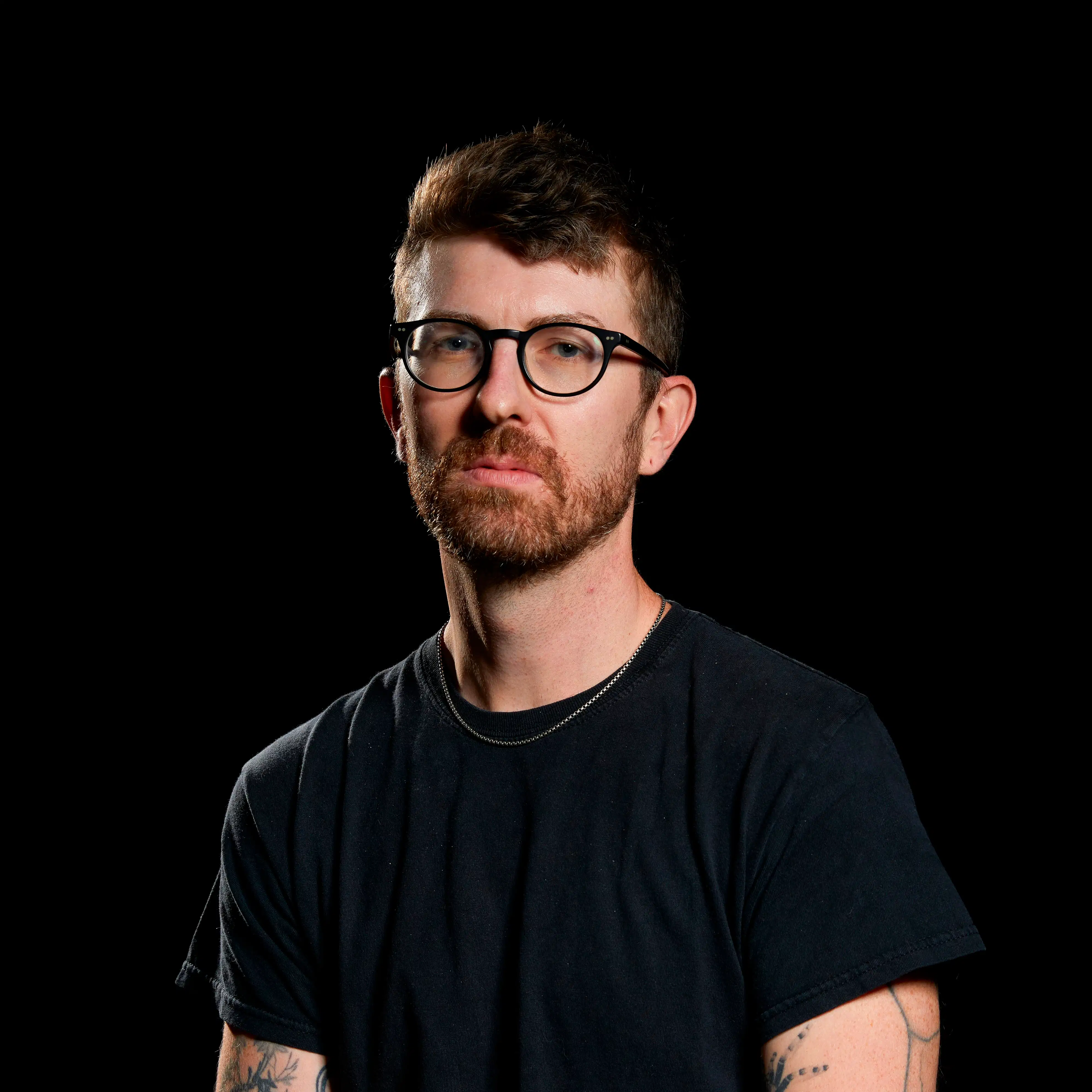 portrait of a man in a studio environment with a dark background wearing a black tshirt. He is wearing glasses and has a silver necklace. 