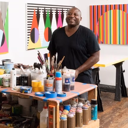 Rico Gatson in a black t-shirt smiles in his studio, surrounded by bold, geometric paintings. 