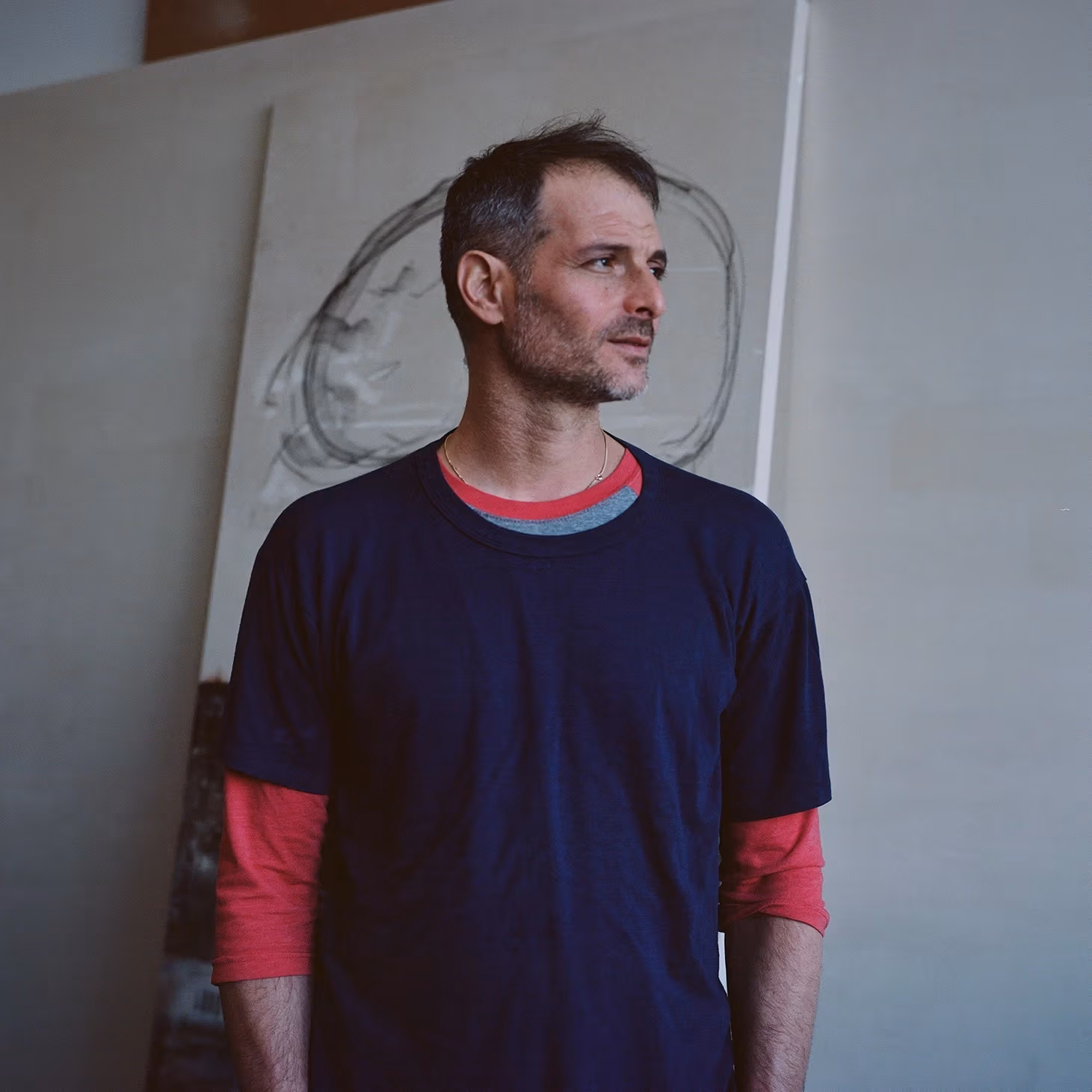 Uri Aran in a navy t-shirt and red long sleeves stands in an art studio, gazing to the side, with a large abstract canvas behind him.