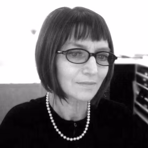 A black and white photo of Debra Klomp Ching, who is looking into the camera with her head slightly turned to the right. She has light skin, short hair, and is wearing a pair of glasses with black frame. She is wearing a black buttoned blouse and a pearl necklace. 