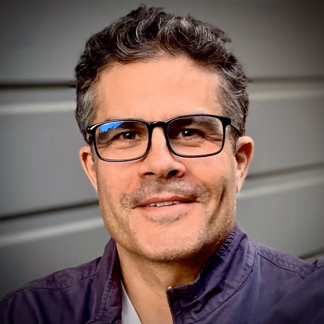Color photo of Mark Tribe showing head and shoulders. He is wearing glasses, smiling, and shown against a gray background. 