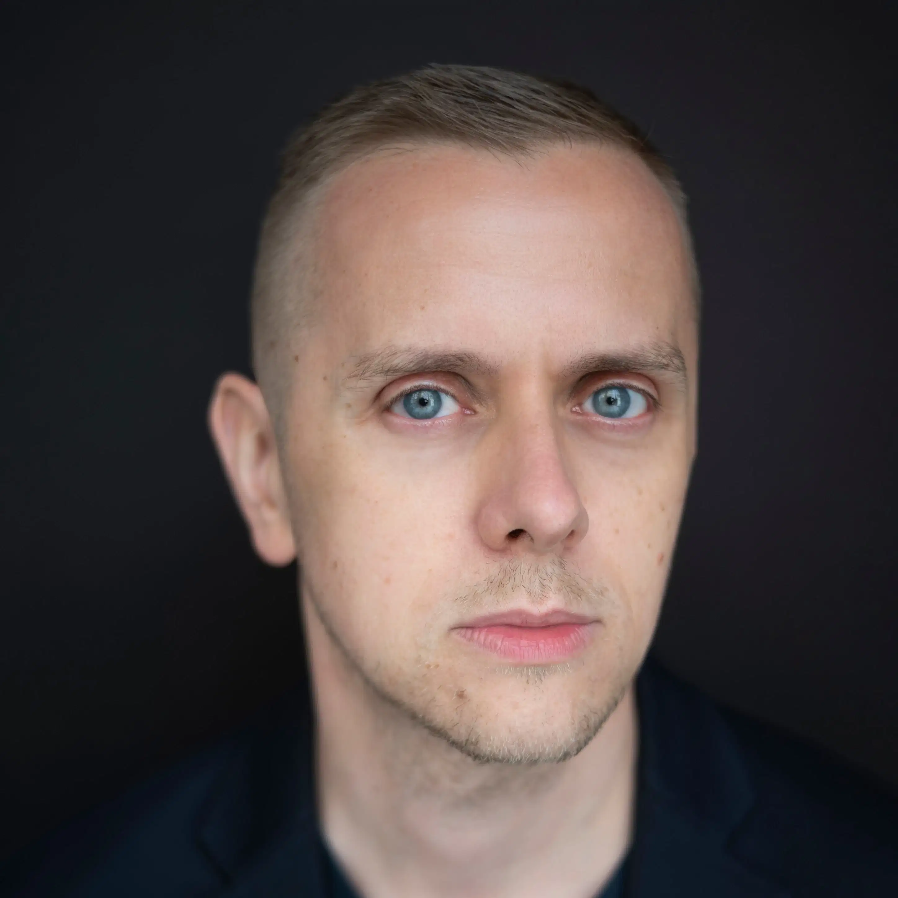 A portrait of Marko Kovacevic looking into the camera against a black background. He has short, blond hair and blond beard. 