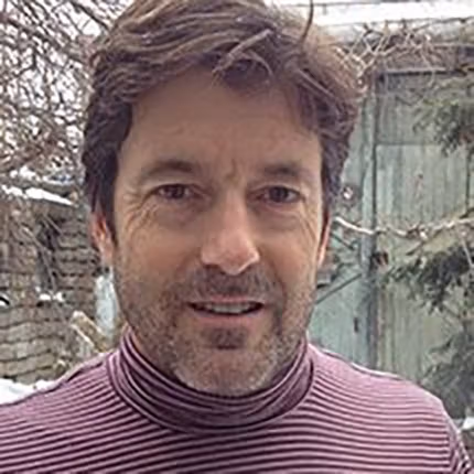 A portrait of Peter Garfield outside in a snowy yeard, wearing a pink and black striped turtleneck sweater