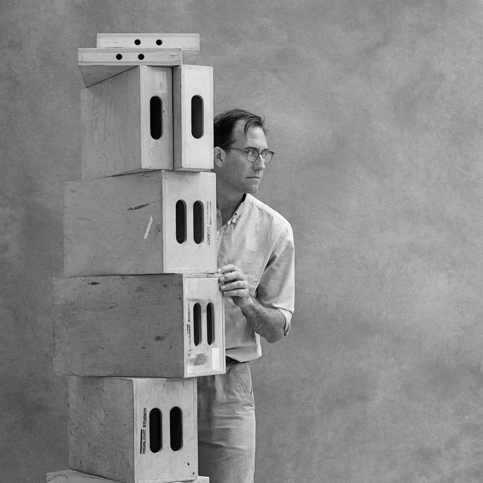 Black and white image of man standing behind a tall stack of studio "apple boxes" looking away. The man is wearing glasses.