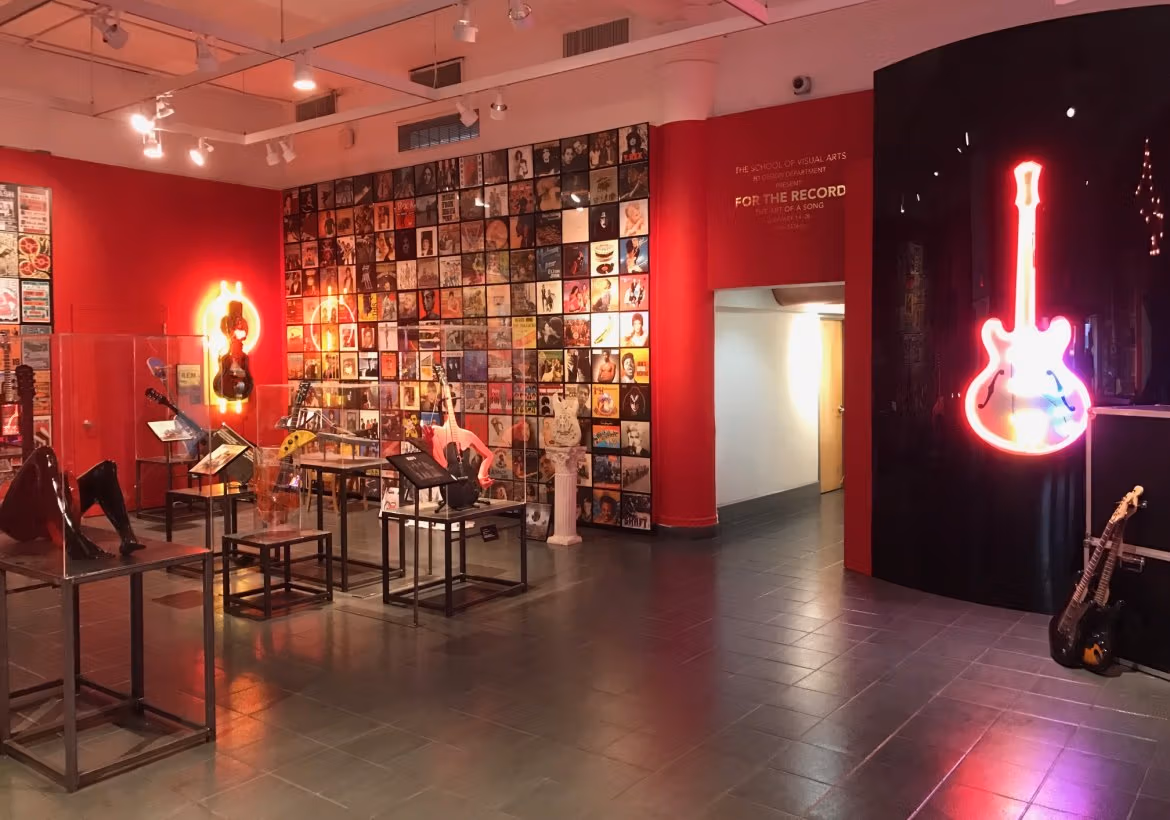 A guitar outlined in neon lights hangs on a wall. Metal stands hold artwork in the center of the room.