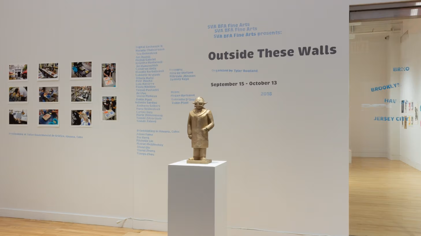 Photograph of external gallery wall with "Outside These Walls" exhibition title, artist names, and printed photographs on the wall. In the center of the room, in front of the wall, is a bronze sculpture of a Viking.