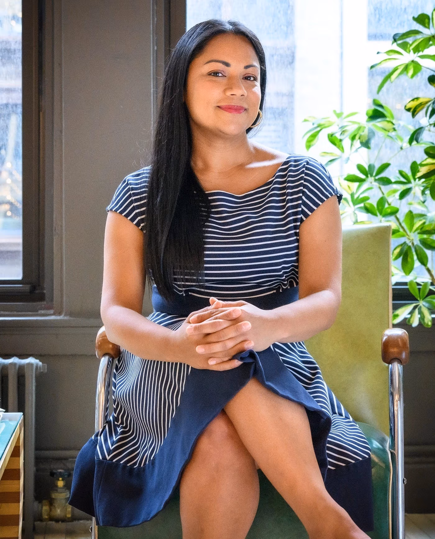 A person with long dark hair wearing a navy and white striped dress sits on a green chair, hands clasped, in a bright room with large windows and a leafy plant.