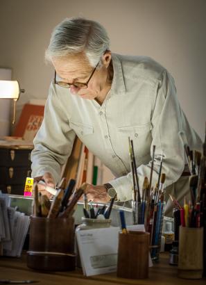 An old man bent over a desk gluing something.