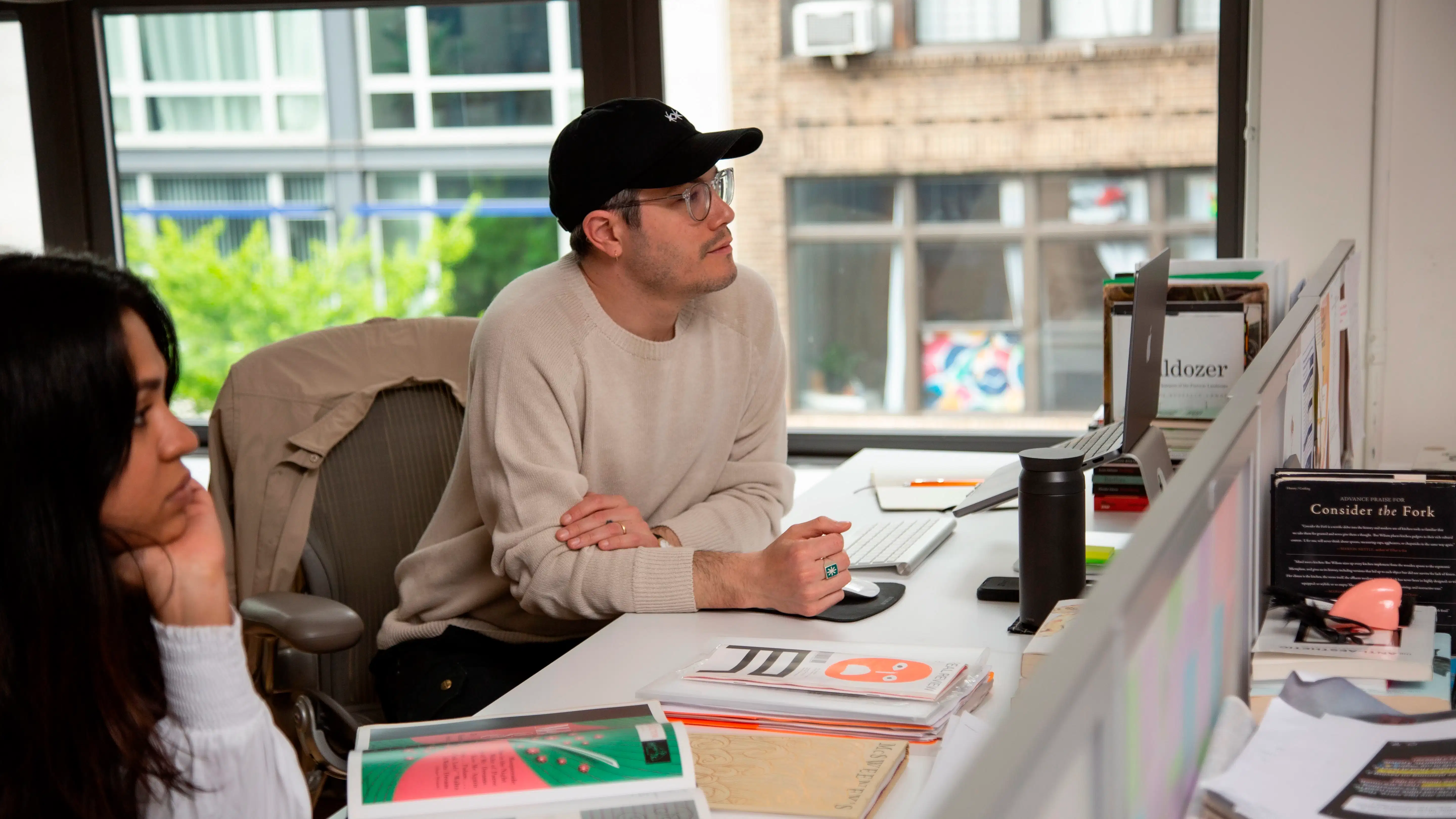 A man in a baseball cap sits at a desk in a bright room next to a woman with long brown hair surrounded by books.