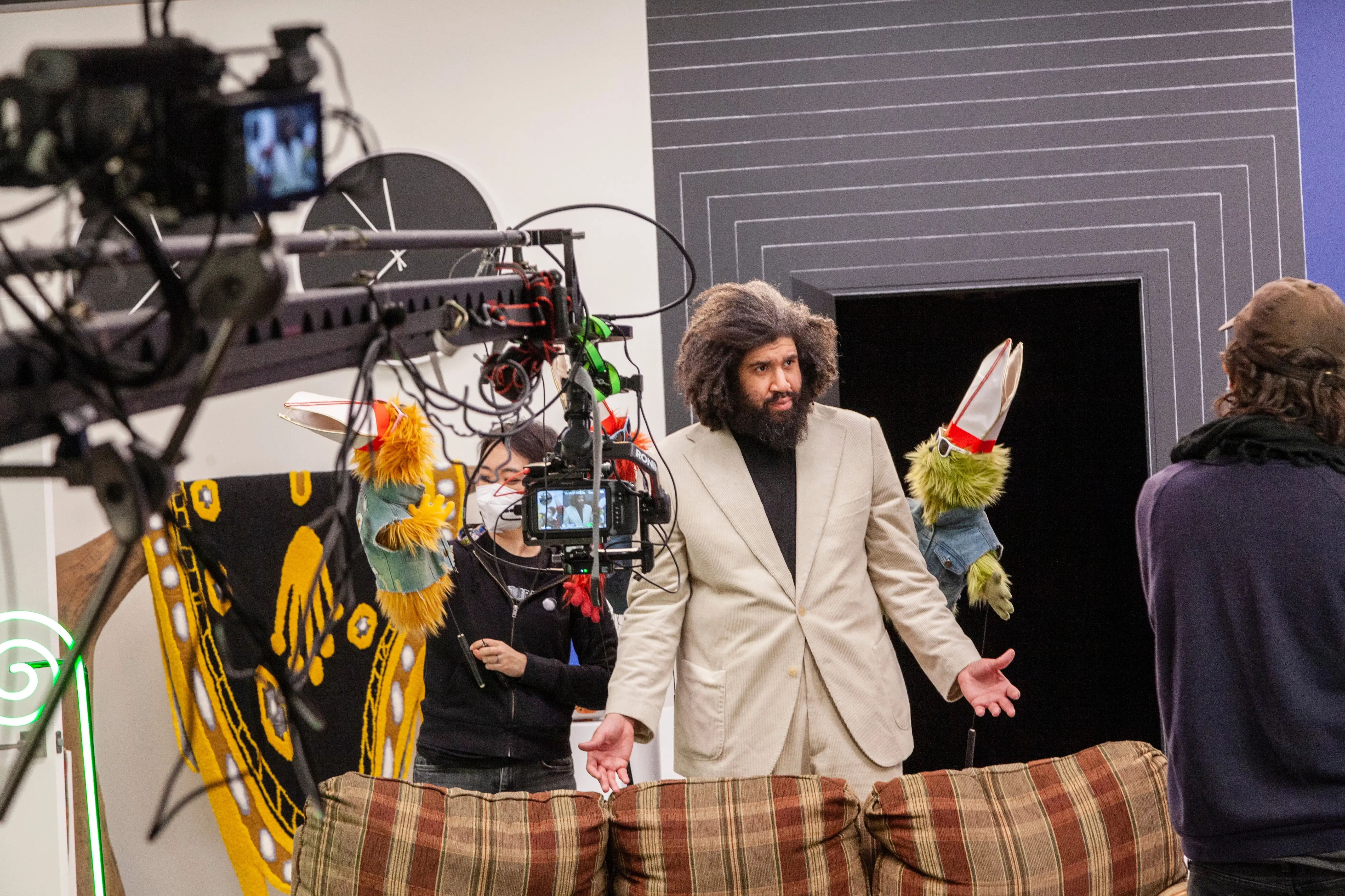 Production still from Jayson Musson's video performance entitled "His History of Art" from 2022. The artist is dressed in a beige suit with a black turtleneck and is performing as "Jay", and standing between two furry puppets, each wearing denim vests, sunglasses and pope's hats. There are production assistants and a camera on an arm in front of him, signaling that this is a behind the scenes shot.