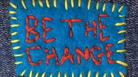 A blue square of fabric with yellow stitching around the borders. The words Be The Change, are stitched in red within the square