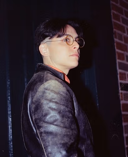 The profile of a dark-haired man wearing a worn leather jacket. The collar of his shirt can be seen and he is also wearing circular glasses.